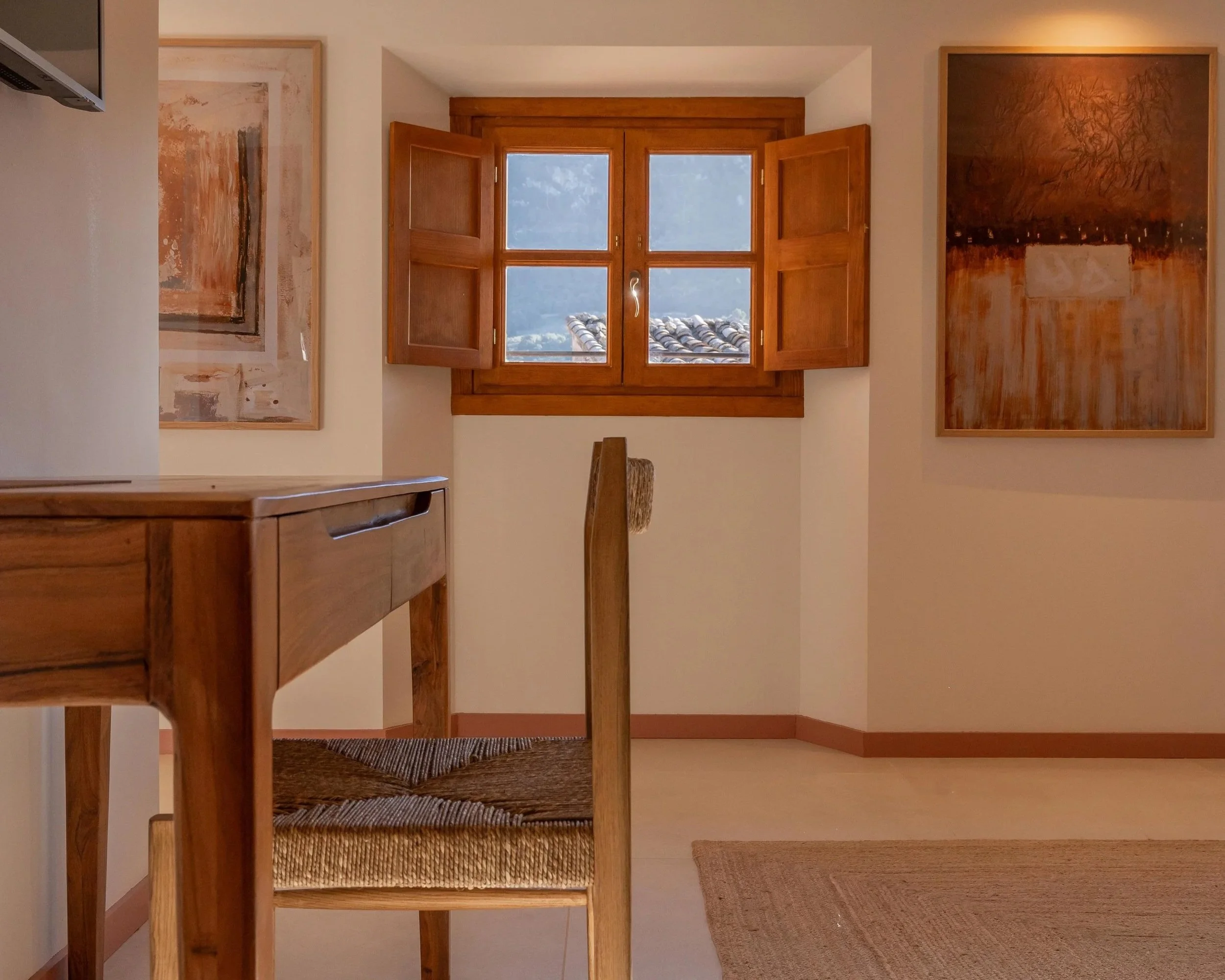 Una habitación con paredes blancas, decorada con dos cuadros abstractos enmarcados en madera, una ventana de madera con contraventanas abiertas, y muebles de madera, incluyendo una mesa y una silla de respaldo alto con respaldo de fibra. Parte de una alfombra en el piso.