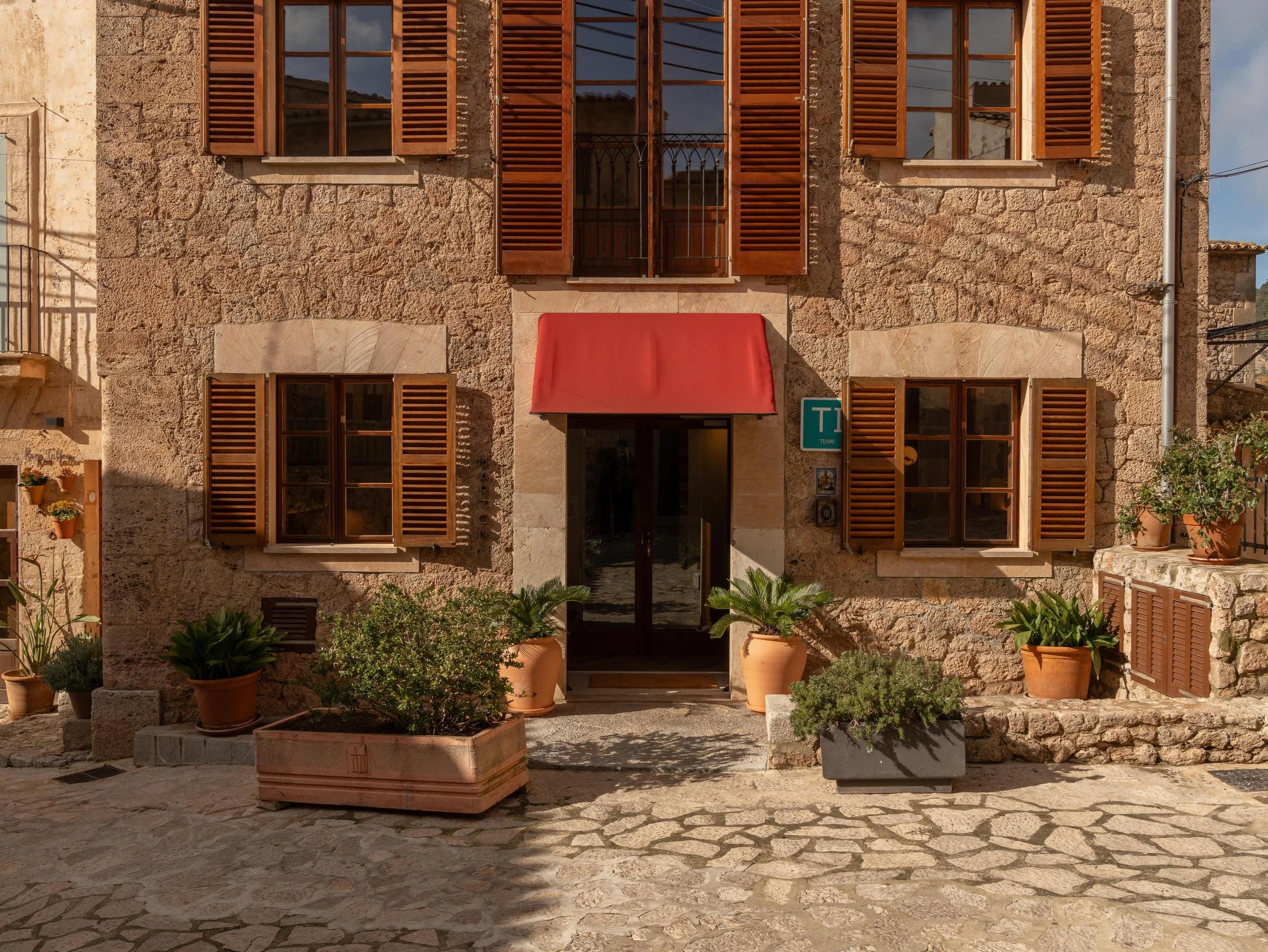 Casa de piedra con ventanas de marcos de madera y persianas. Entrada principal con una cortina roja y varias plantas en macetas a los lados.