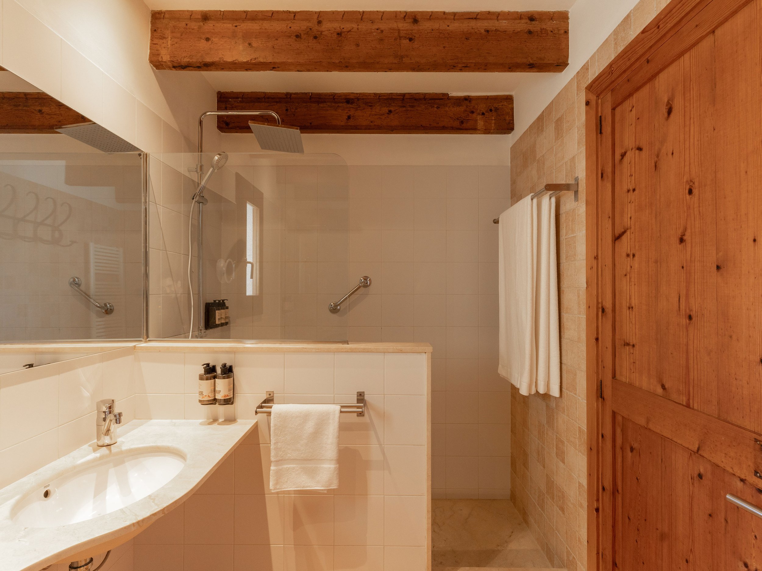 Baño con pared de azulejos blancos, ducha con puerta de cristal, lavabo con lavabo y jabón, toallas blancas en riel, techo con vigas de madera, puerta de madera cerrada.