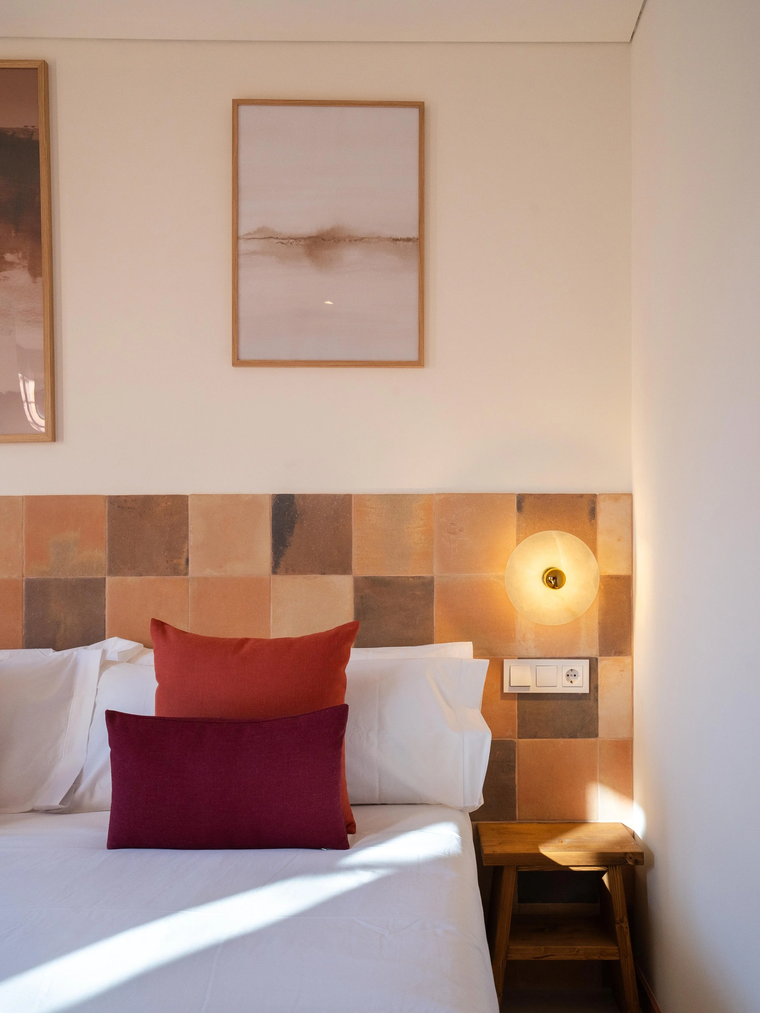 Cama en una habitación moderna con cabecera de azulejos de colores terracota, dos almohadas en tonos rojos y una pared con cuadros y luz de pared
