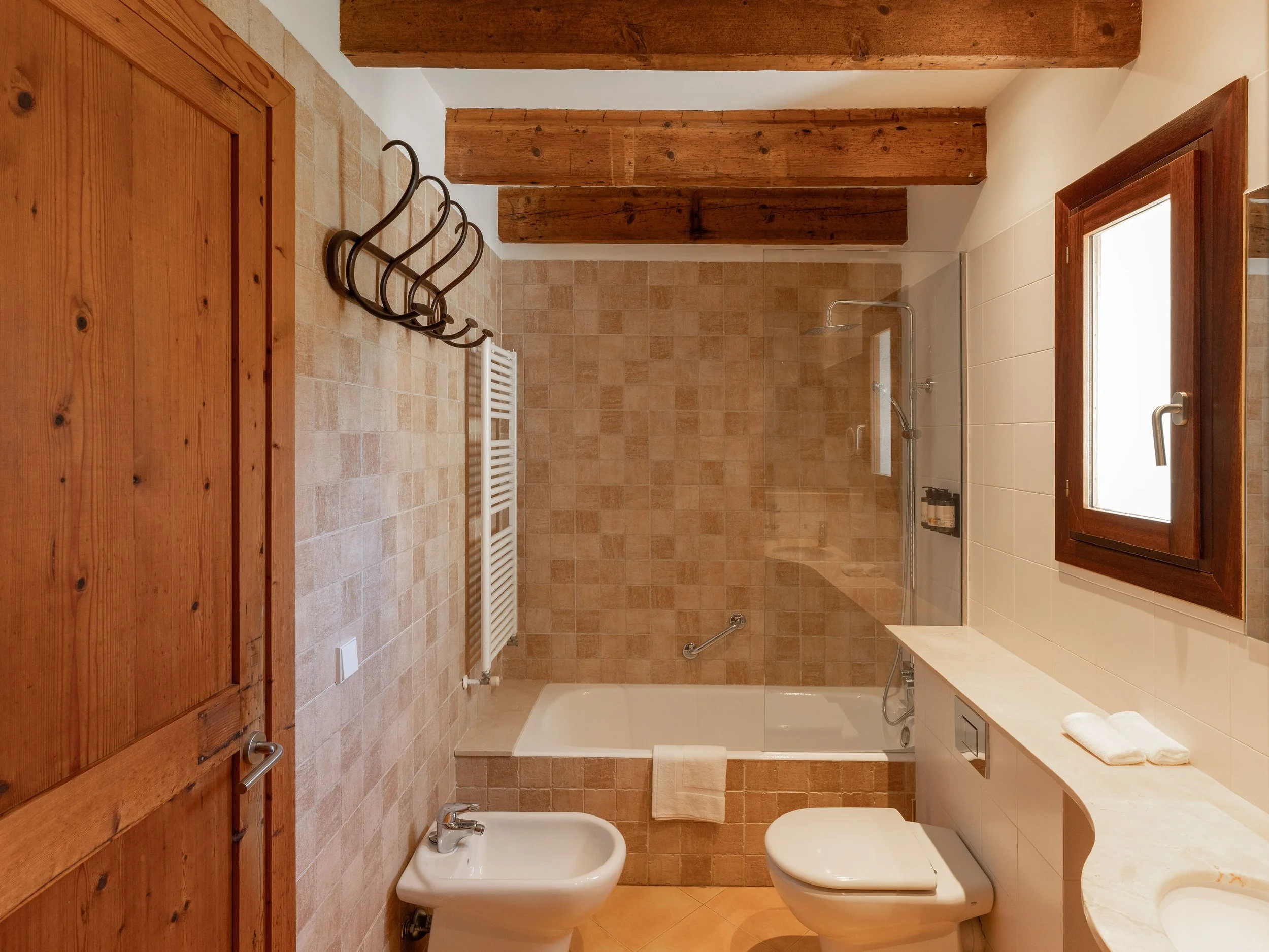 Baño con paredes de azulejo beige, ducha con vidrio y accesorios metálicos, inodoro, bidé, ventana de madera con cristal, armario de madera, toallas y accesorios de baño.