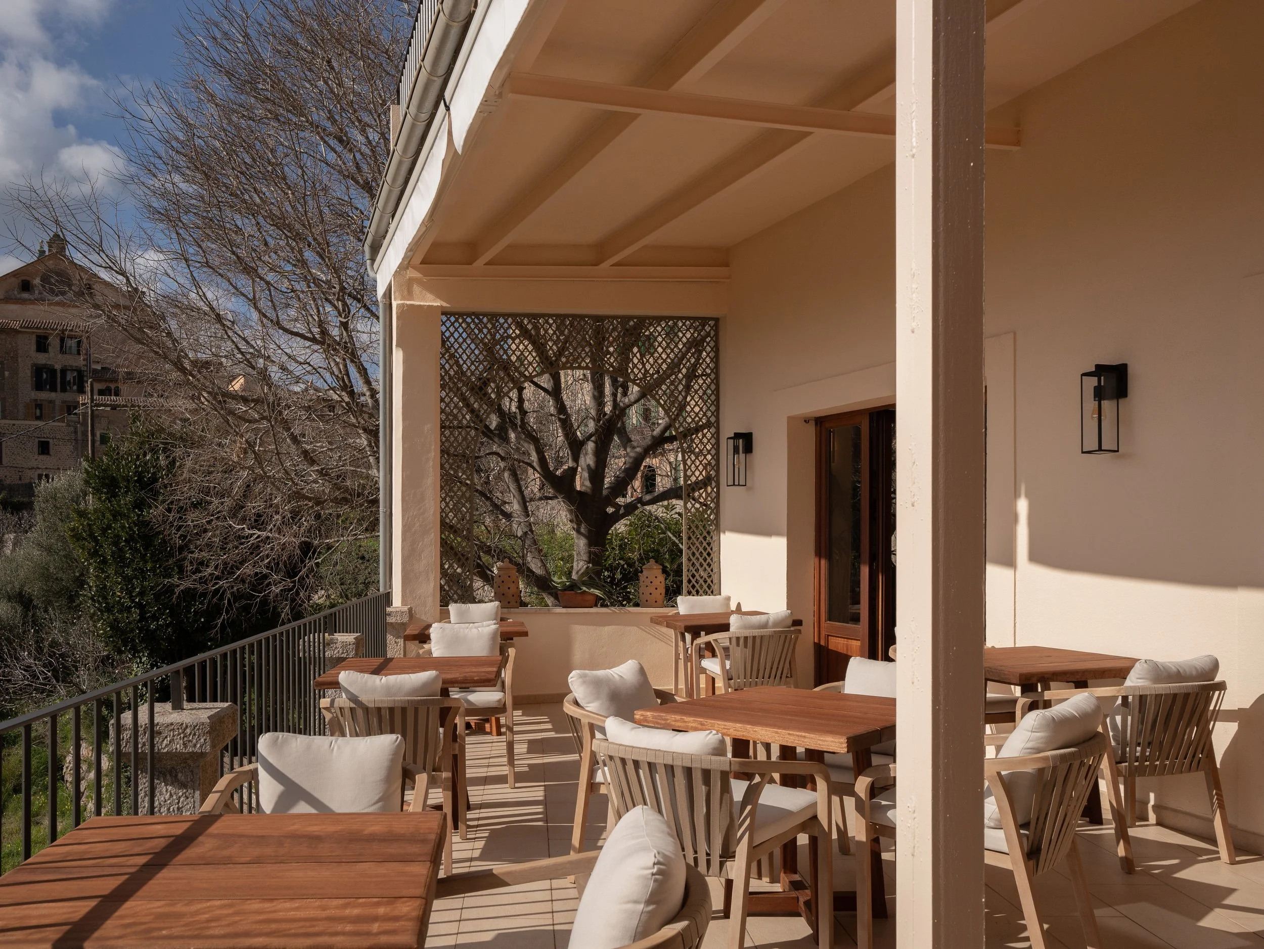 Terraza con mesas de madera y sillas con cojines blancos, árboles desnudos y edificio en el fondo, en un día soleado.
