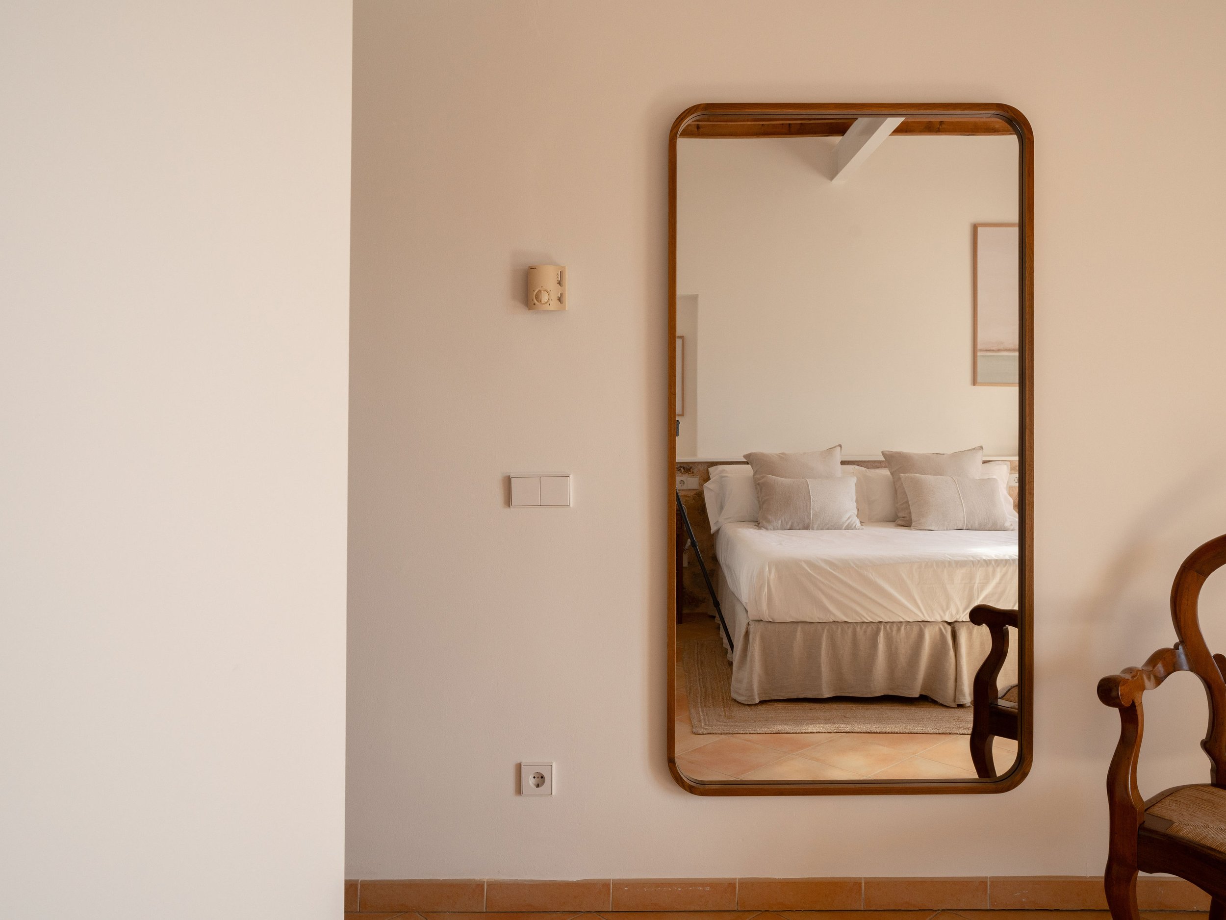 Espejo en la pared que refleja una cama con almohadas en una habitación. La habitación tiene paredes blancas y cuadros en la pared.