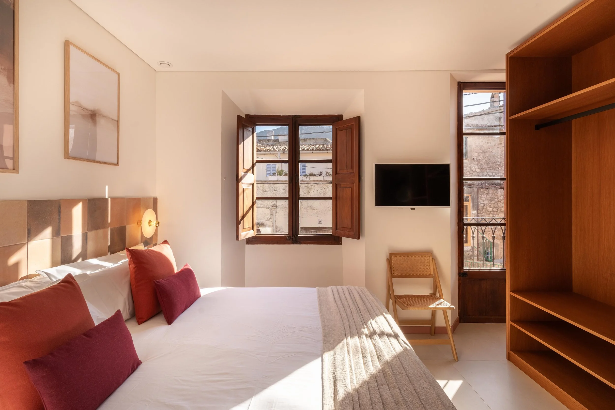 Habitación con cama, almohadas color terracota y vino, ventanas de madera abiertas, televisión, mesa y estantería de madera, luz natural