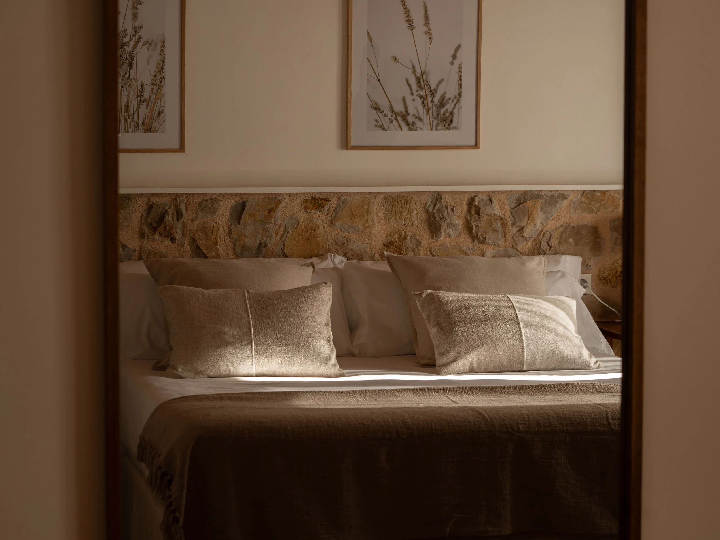 Cama con almohadas beige y un cabecero de piedra, decorada con cuadros de plantas.