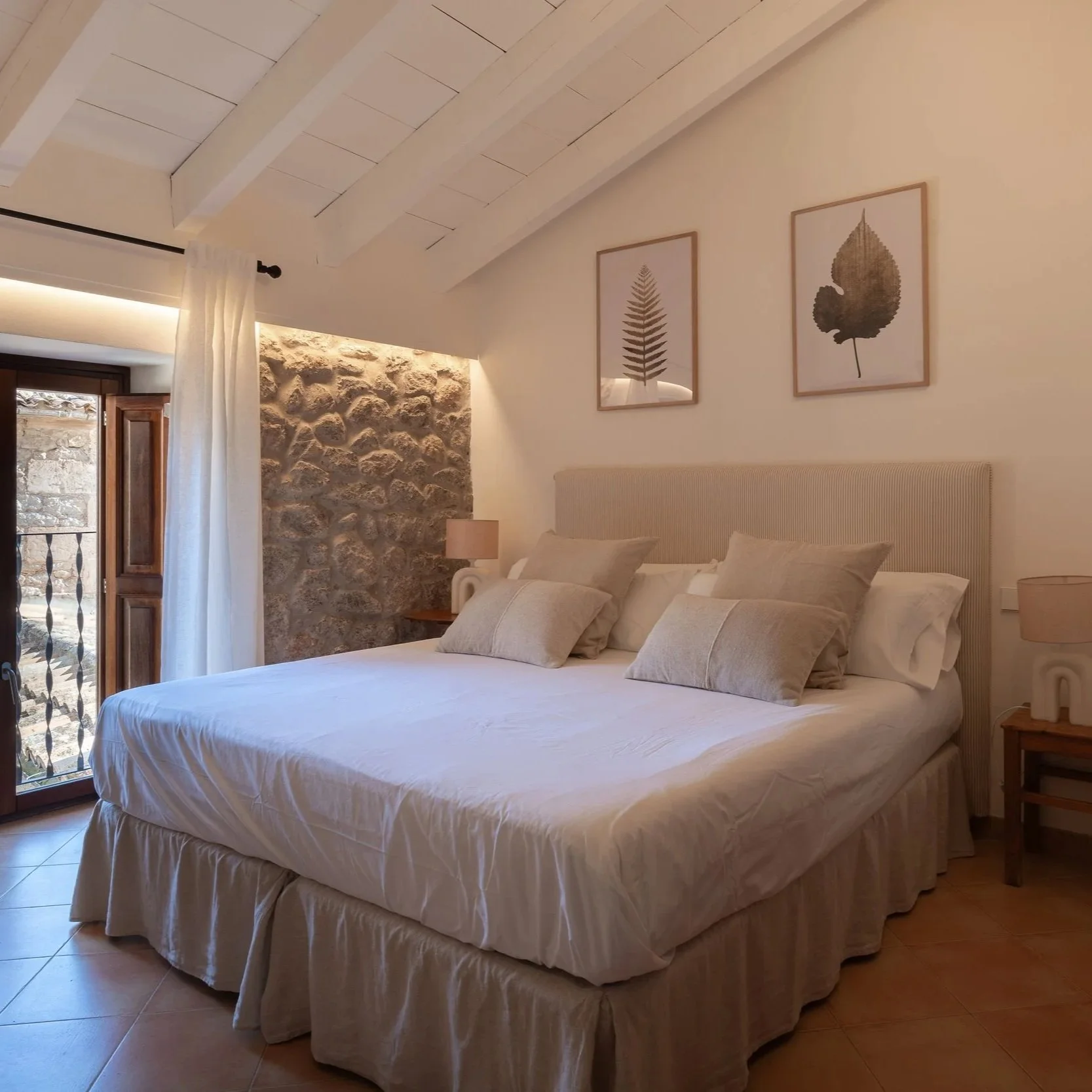 Habitación con cama grande, cabecera acolchada, almohadas beige, paredes blancas, cuadros con hojas, lámparas de noche, balcón con puertas de madera y cortinas blancas, pared de piedra decorativa