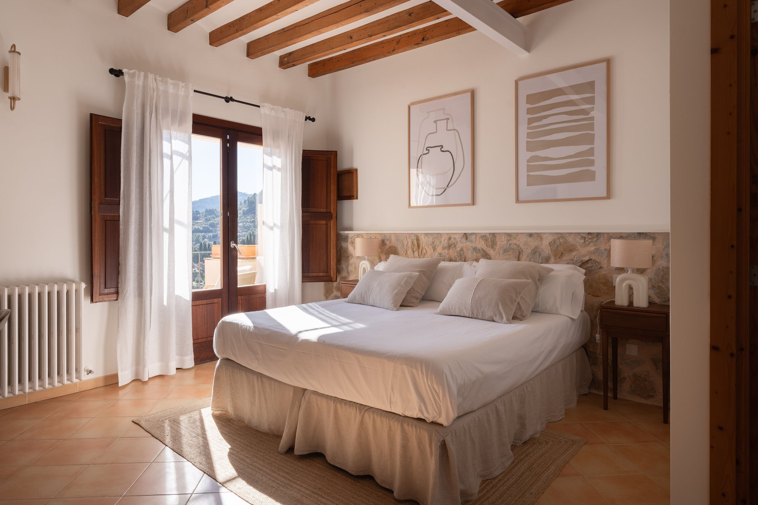 Habitación con cama grande, paredes blancas, cortinas blancas, ventanas de madera con vista a montañas, cuadros abstractos en la pared, lámparas de mesa, suelo de baldosas de color terracota, techo de madera con vigas visibles.
