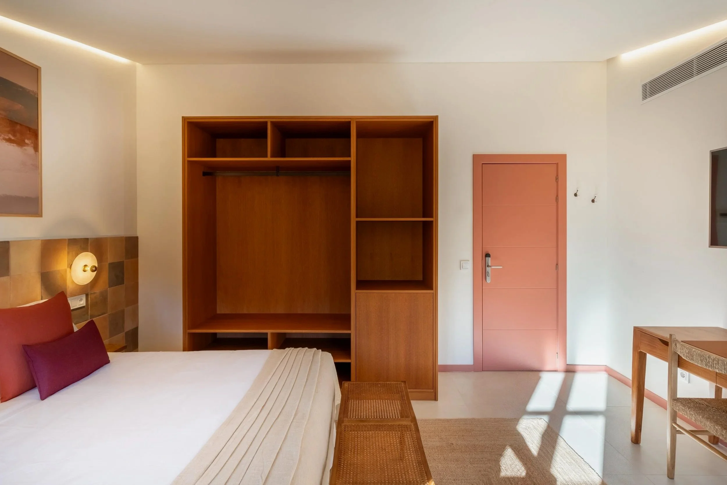 Habitación moderna con cama, cabecera de madera con color en tonos terracota, mesita de noche con lámpara, armario de madera, pared blanca y puerta rosada, decoraciones sencillas y natural luz.
