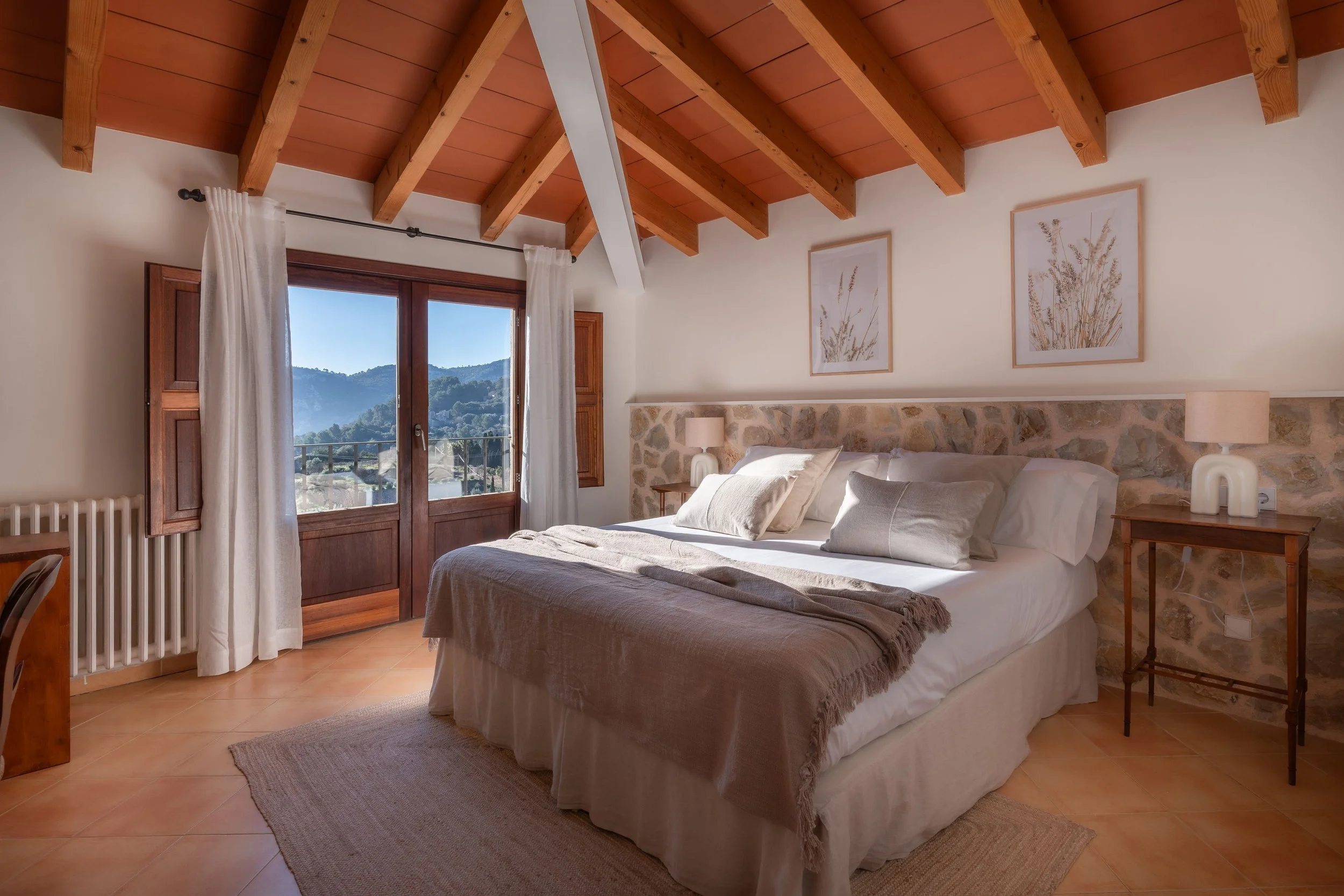 Habitación con cama de matrimonio, paredes de piedra, ventanas con vista a montañas, techos de madera y decoraciones neutras.