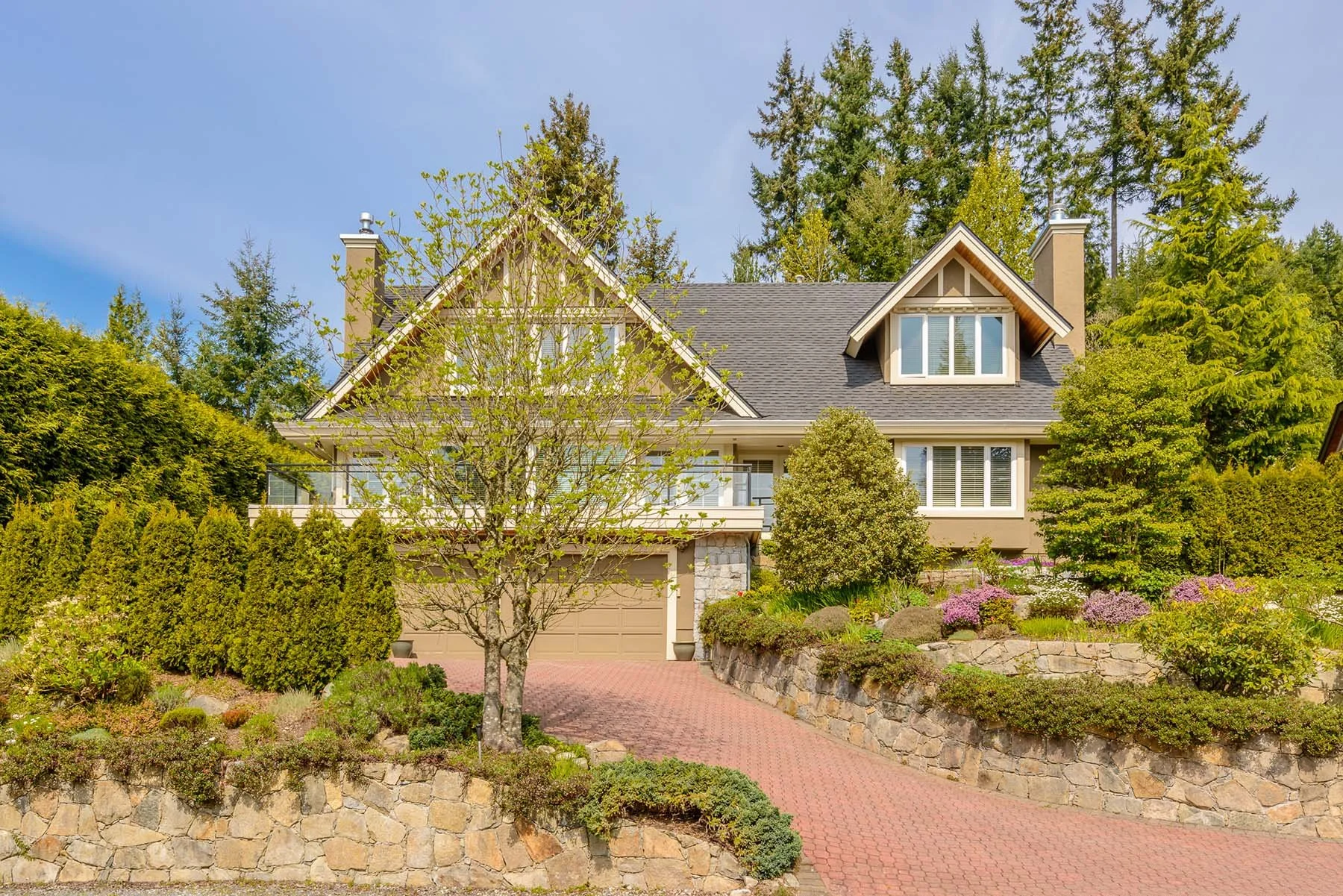 A large house with beige exterior walls, a dark gray roof, and multiple windows, surrounded by a lush green garden with trees and shrubs, and a stone driveway leading to a garage.