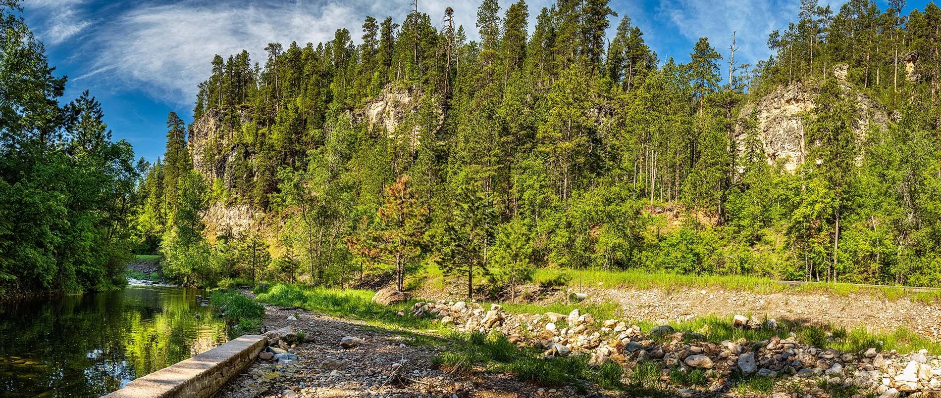 A scenic landscape with a river on the left, lush green trees, and a rocky hillside under a partly cloudy sky.