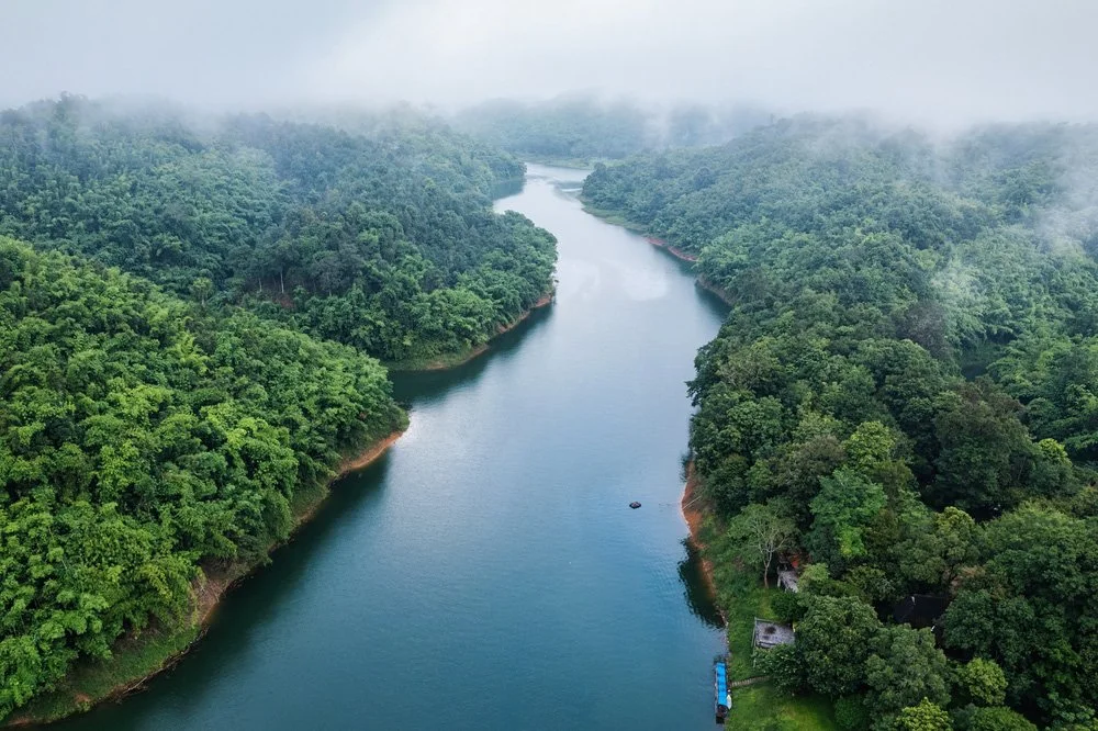 Aerial view of a winding river flowing through dense green forested hills with misty clouds overhead.