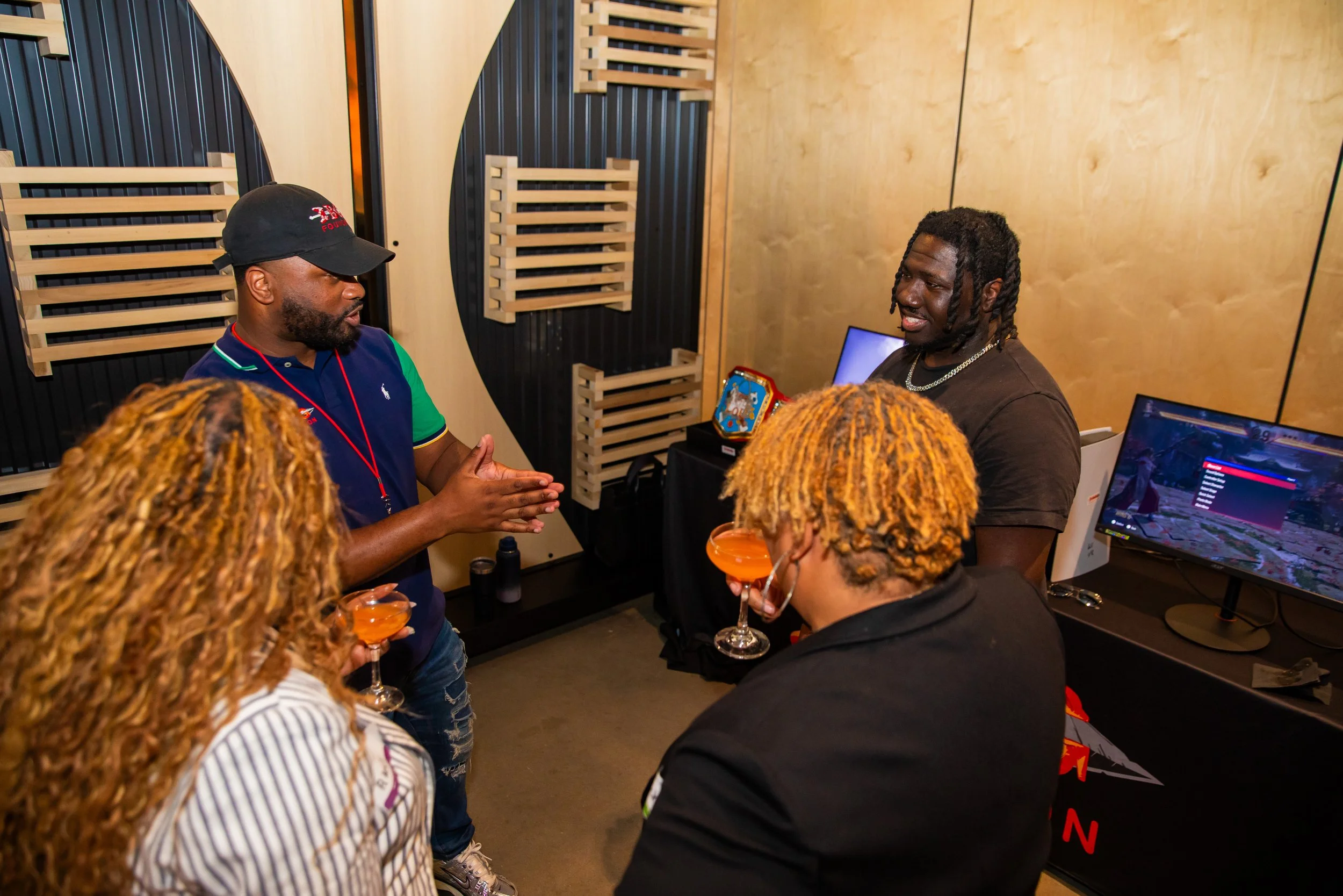 A group of people socializing indoors, with two men at the center engaged in conversation, one wearing a black cap and the other with dreadlocks. Some individuals are holding drinks, and there are computer screens and a trophy in the background.