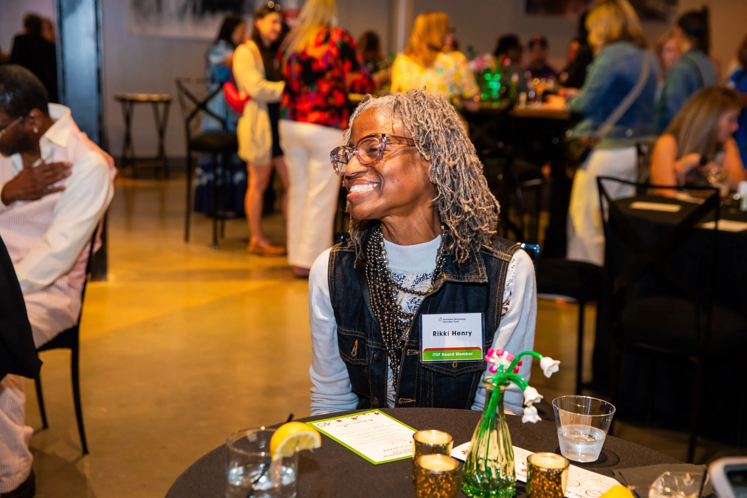 A woman with gray dreadlocks and glasses smiling at a celebration or event, seated at a table with food and drinks, wearing a name tag that reads 'Rikki Henry' and indicating she is an ITEF Board Member.