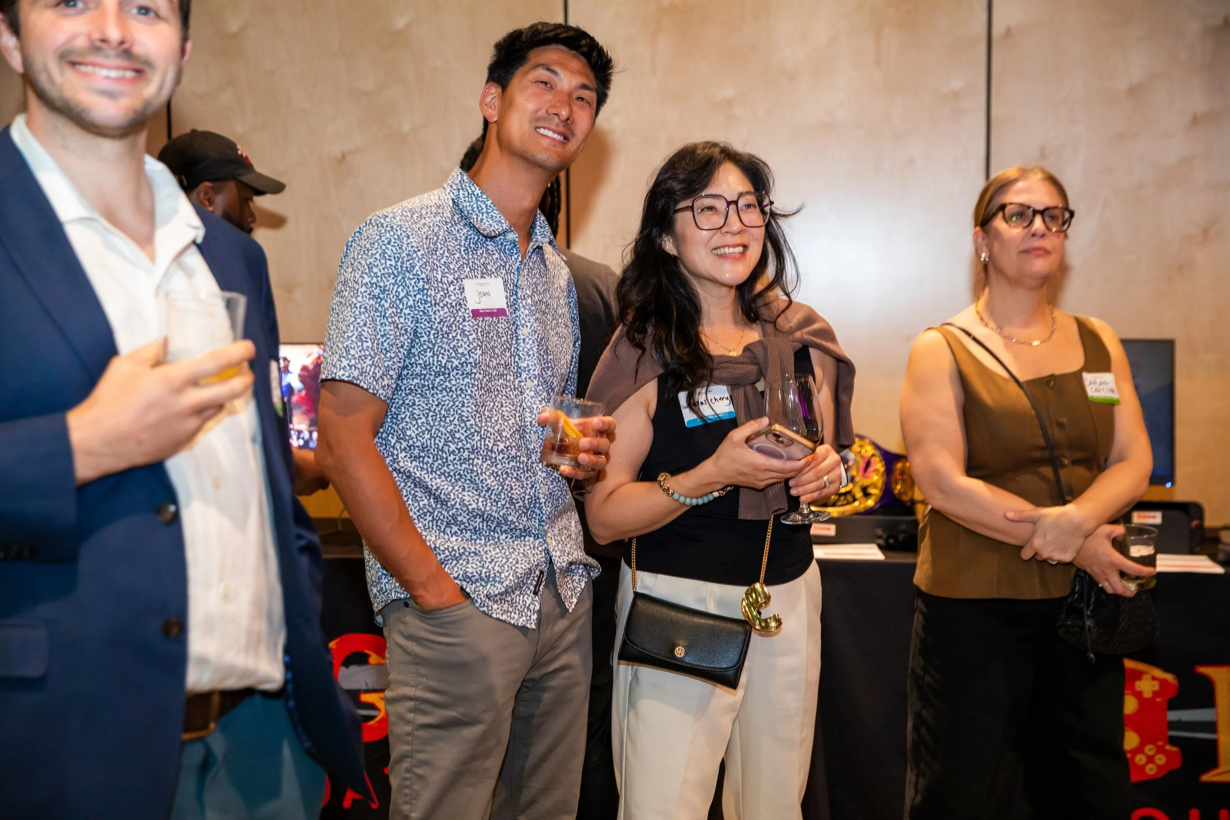Group of diverse people standing together at an event, smiling and holding drinks, with some wearing name tags.