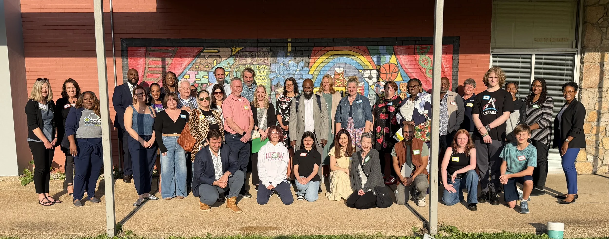 Group of diverse people posing outside in front of a colorful mural, some wearing name tags and casual clothing.
