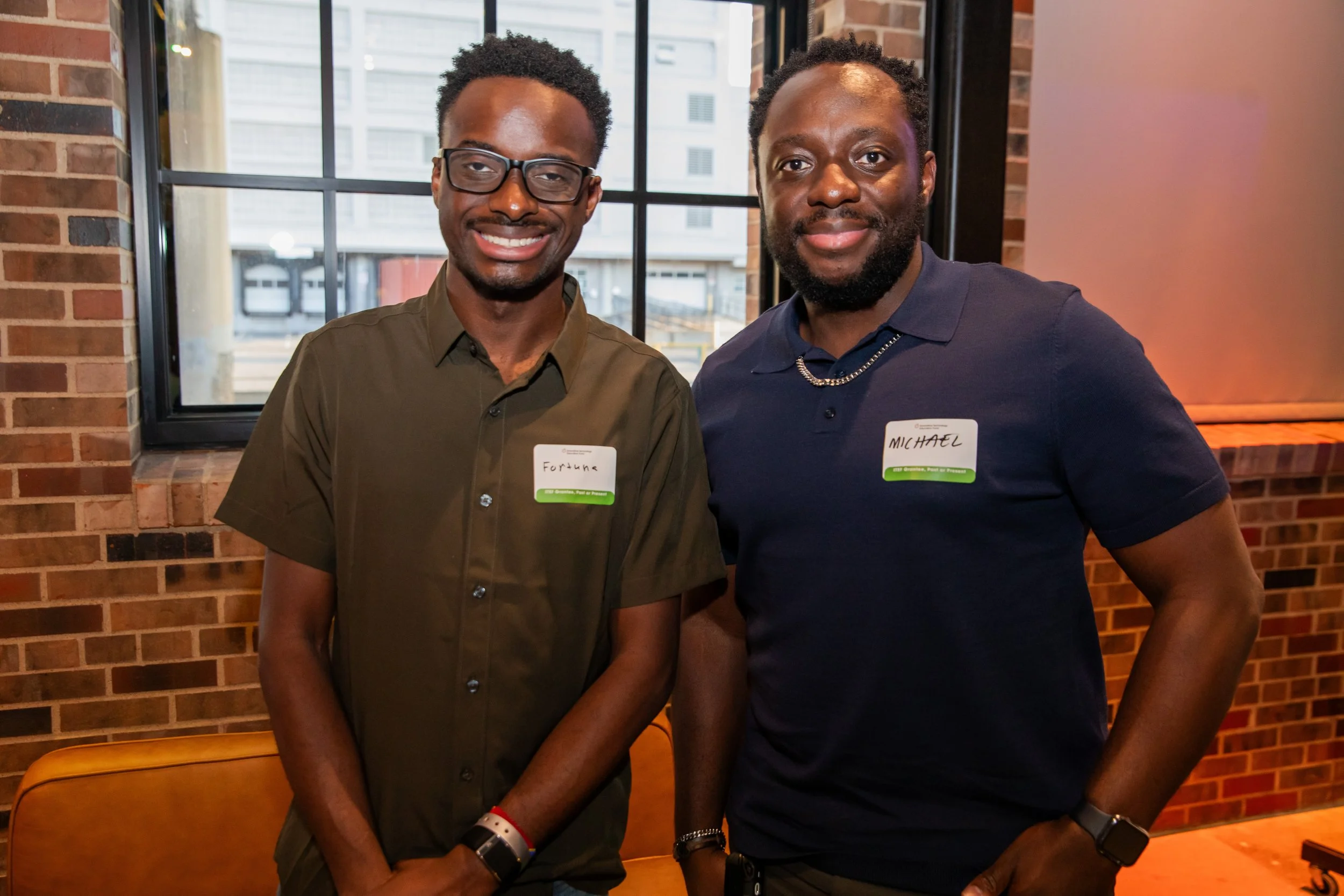 Two men standing side by side smiling at the camera in a room with a brick wall and large windows. One man is wearing glasses and a brown shirt, with a name tag that says 'Fortune.' The other man has a beard, is wearing a navy shirt and a chain neckl