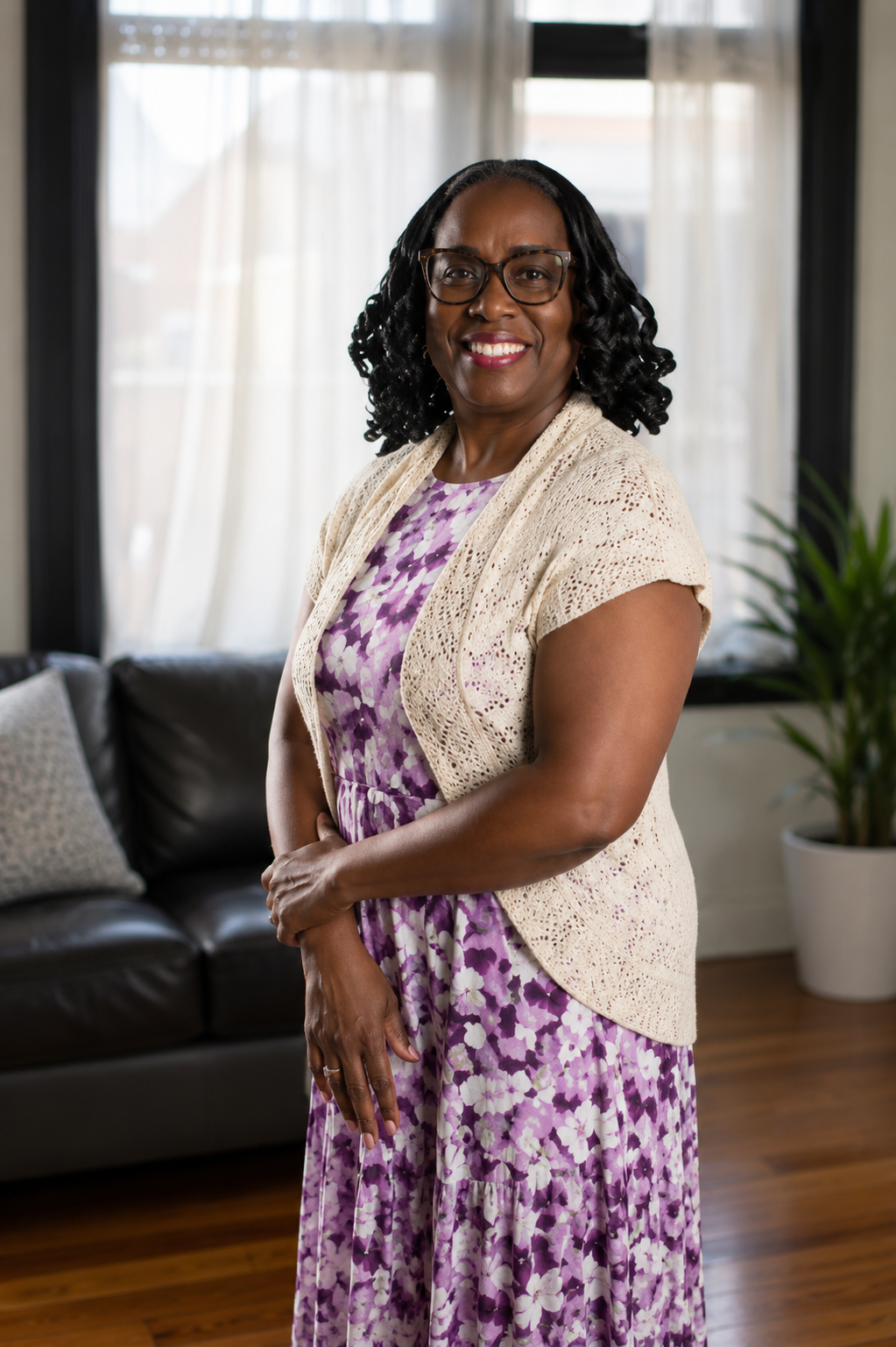 A smiling African American woman with glasses, black curly hair, wearing a purple and white floral dress and a cream cardigan, standing in a living room with large windows, curtains, a dark sofa, cushions, and a potted plant.