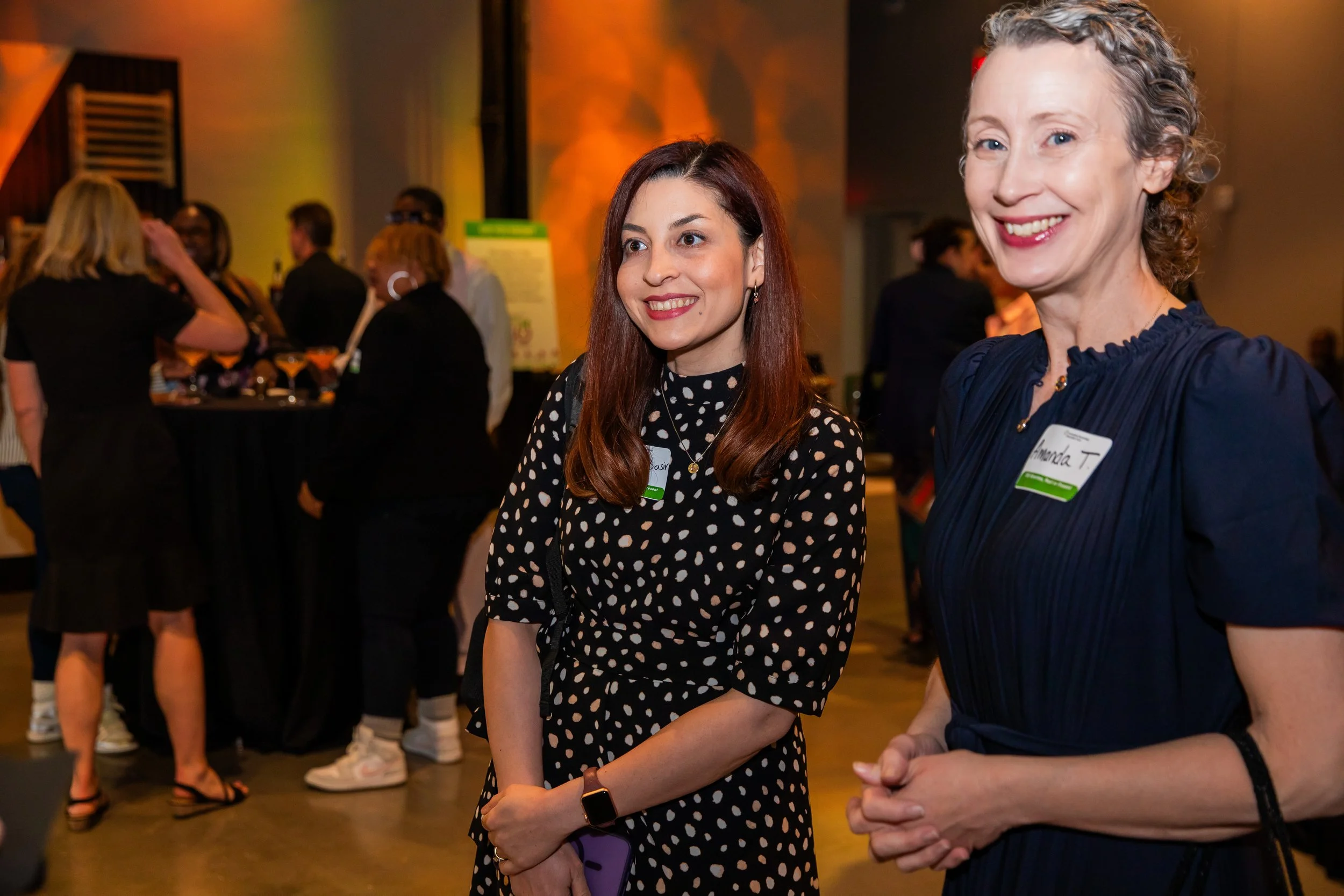 Two women attending an indoor event, standing close together and smiling, with other people behind them. The woman on the left has long red hair and wears a black dress with white polka dots. The woman on the right has short curly gray hair and wears