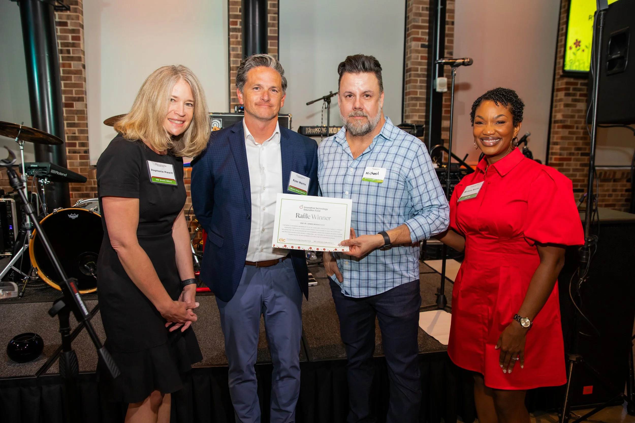 Four people standing together at an event, one man is holding a certificate, all are smiling. Background includes musical instruments and audio equipment, with brick wall decor.
