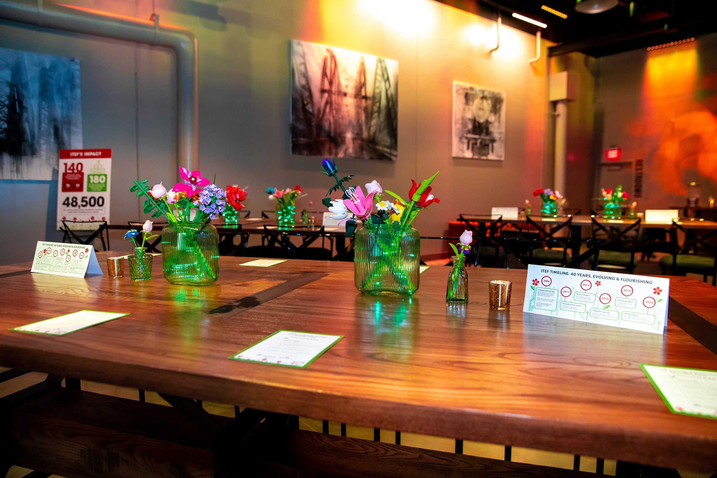An exhibition space with tables showcasing colorful flower arrangements in green glass vases. The space has artwork on the walls and informational placards on the tables. The lighting creates a warm, vibrant atmosphere.