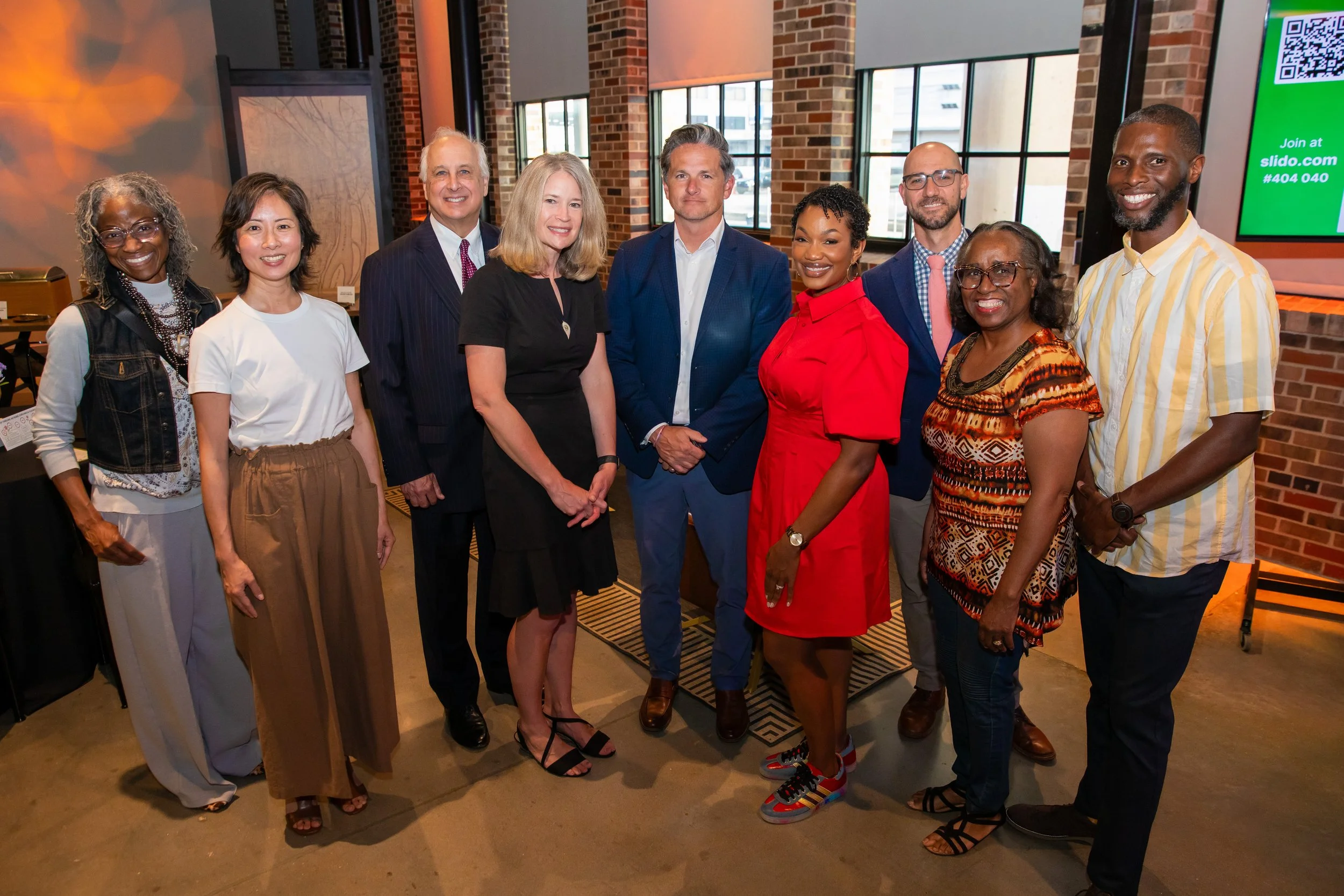 A diverse group of ten people standing together indoors, smiling for the photo. The background features brick walls, large windows, and a digital screen with a QR code.