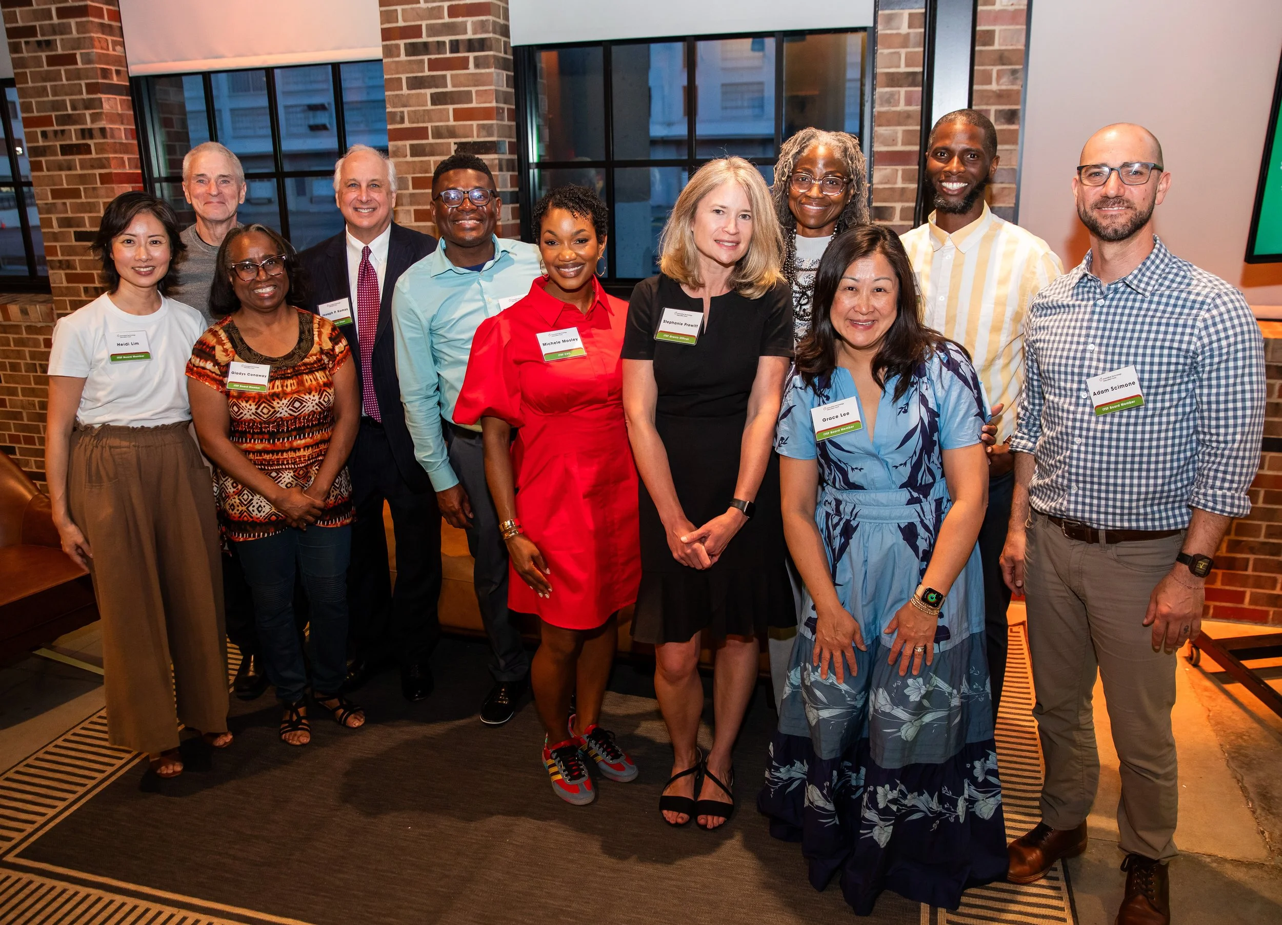 A diverse group of eleven people posing for a photo in an indoor setting with brick walls and large windows. They are smiling and wearing name tags.