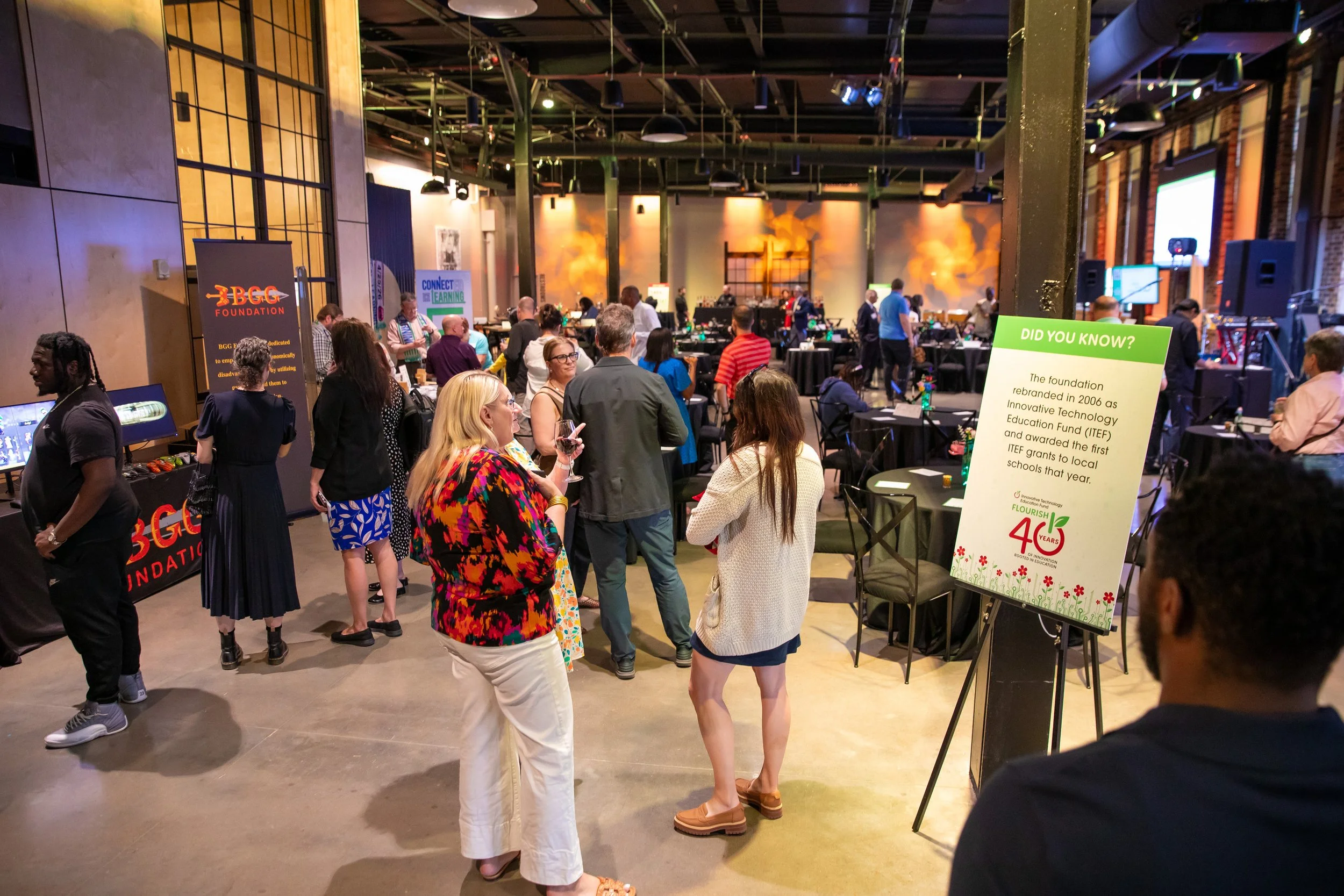 A busy indoor event with numerous people socializing and walking around, featuring various informational displays, tables, and a stage area at the back with warm lighting and brick walls.