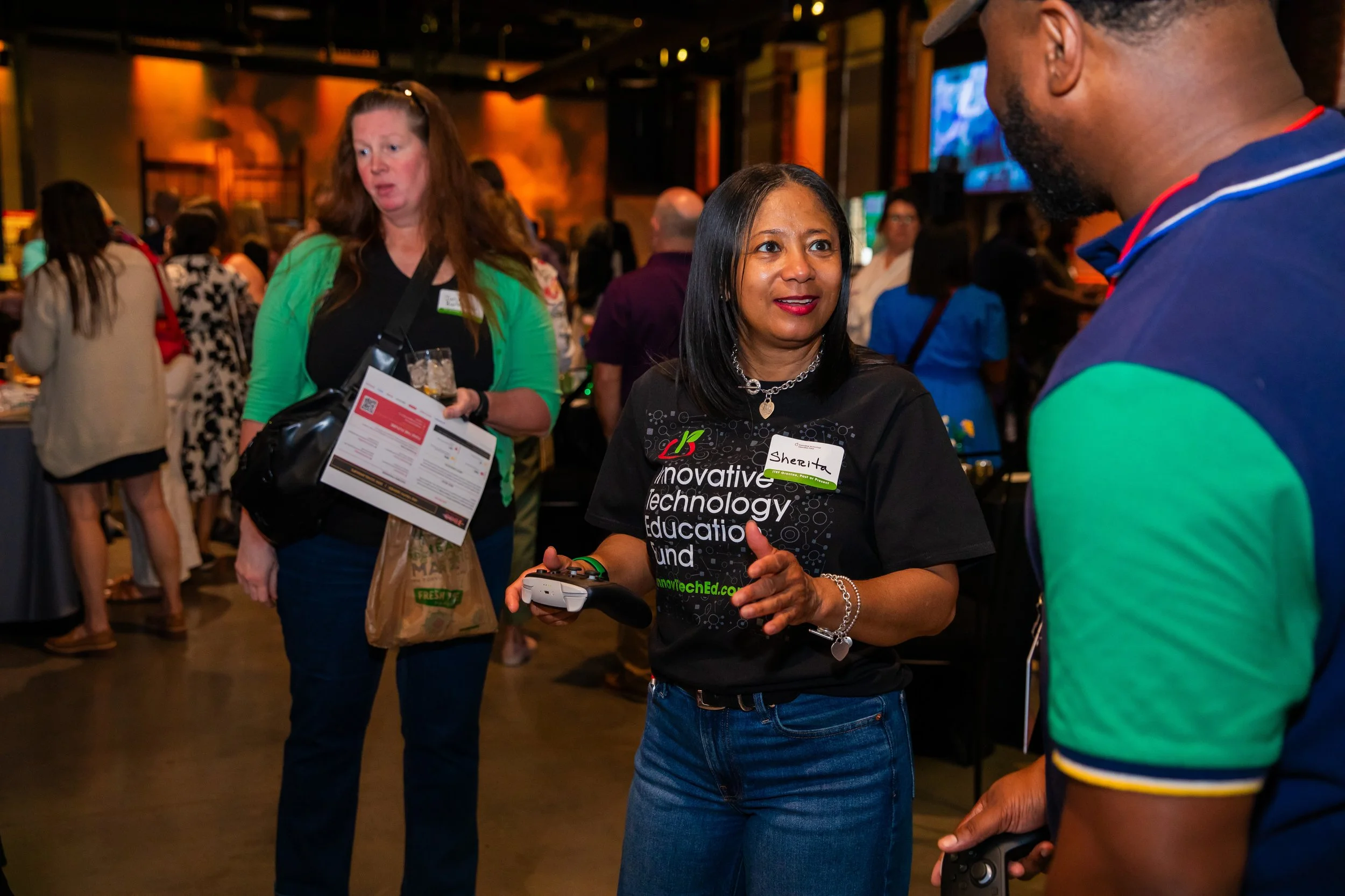 Two women and one man in conversation at an indoor event. One woman wearing a black shirt with white text, the other woman in a green cardigan, and a man in a colorful shirt. Background features other people, some holding drinks, and a large televisi