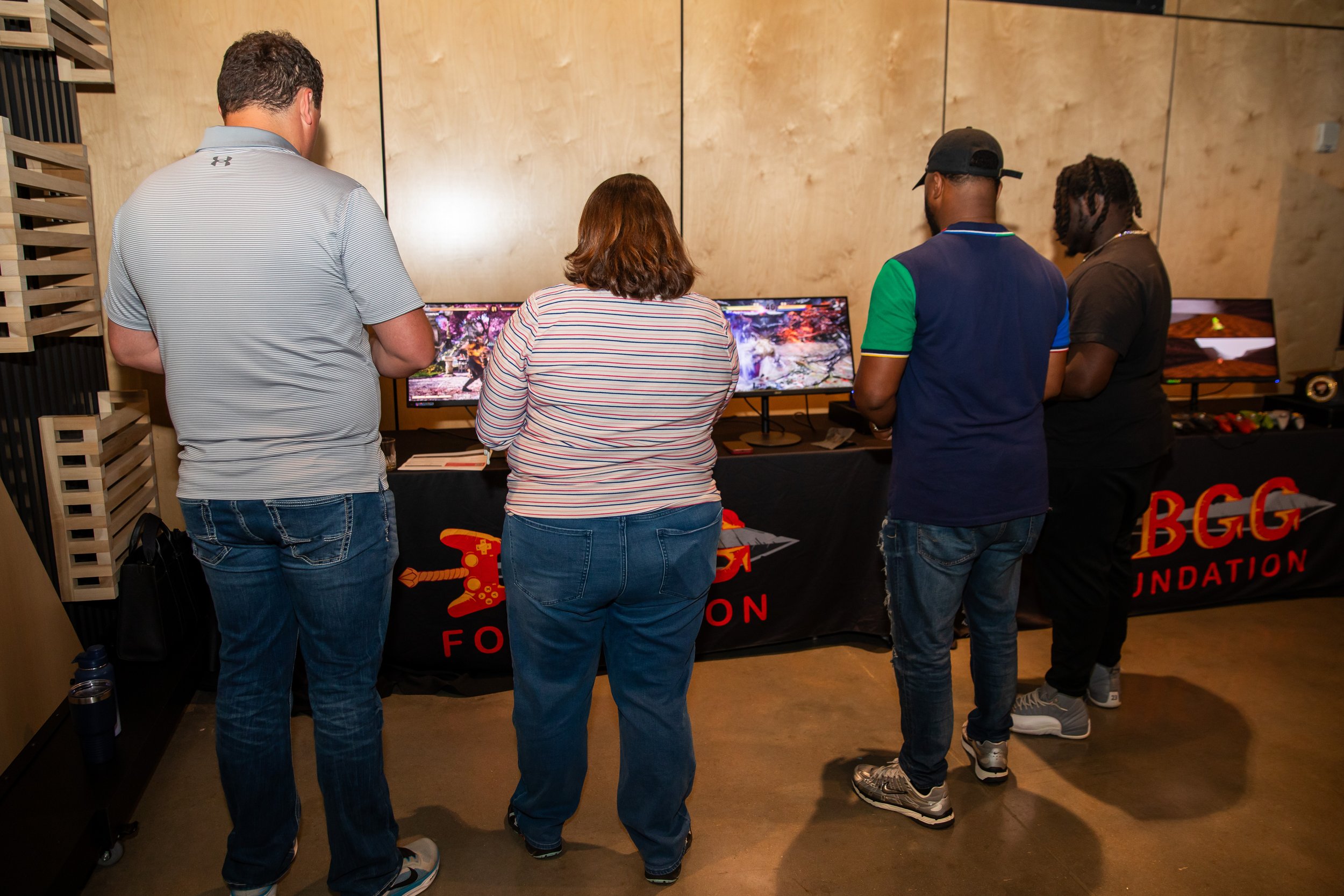 Four people are standing with their backs facing a row of computer monitors at an indoor event. The monitors display a video game scene. There is a table underneath the monitors with a black tablecloth and a red and yellow logo that reads 'BGC Founda