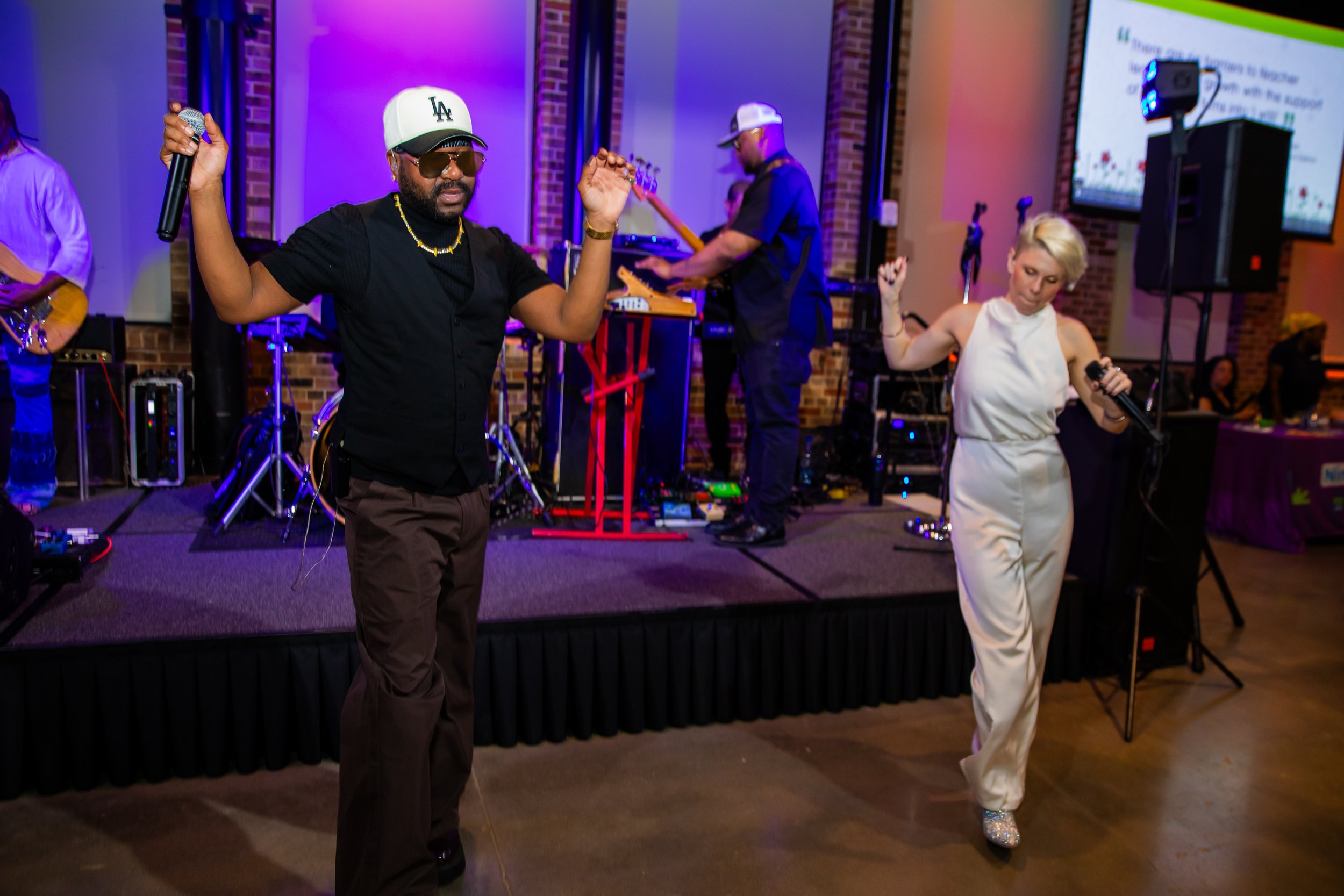 A man wearing sunglasses, a white LA cap, and gold jewelry is dancing with a woman in a white outfit and high heels with sparkles; both are holding microphones at a live music event inside a venue with brick walls and a large screen.