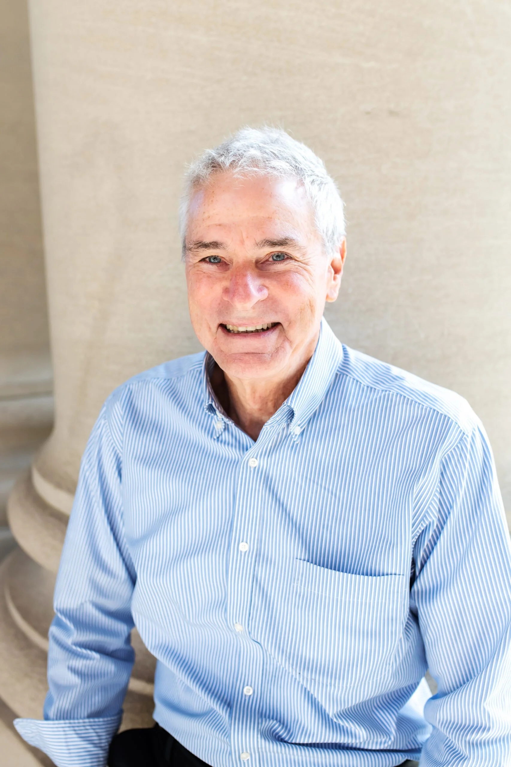 A smiling older man with short gray hair, blue eyes, wearing a light blue and white striped dress shirt, sitting on a beige staircase, with a plain beige wall in the background.