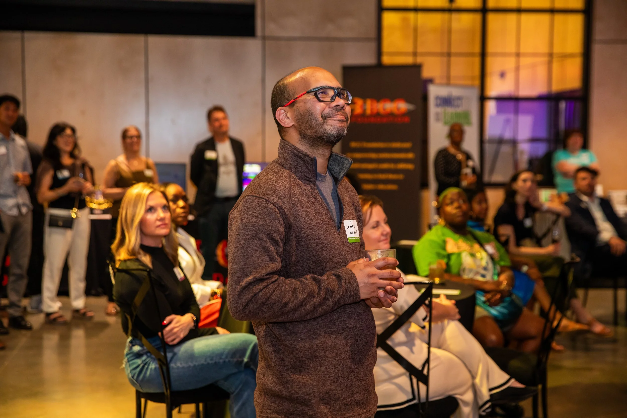 A man with glasses and a brown jacket holding a drink at a conference or event with several seated and standing attendees in the background.