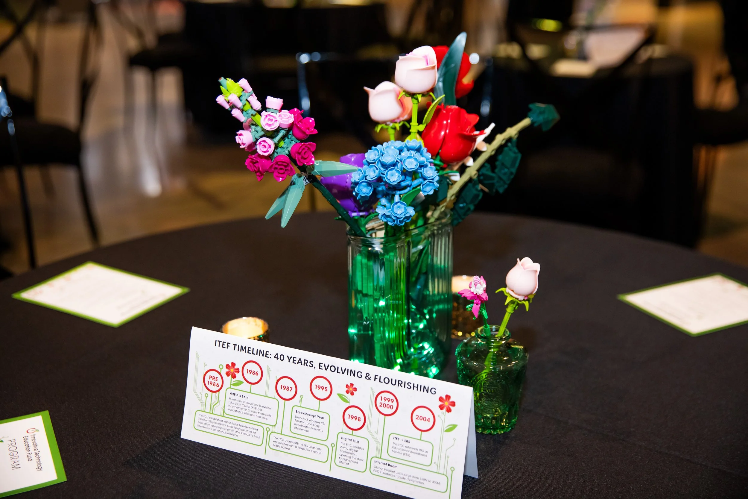 A glass vase with LED lights inside holds a colorful arrangement of toy flowers made from building blocks. There is a small glass container neighboring the main vase with similar toy flowers. The background shows chairs and a table, suggesting a disp