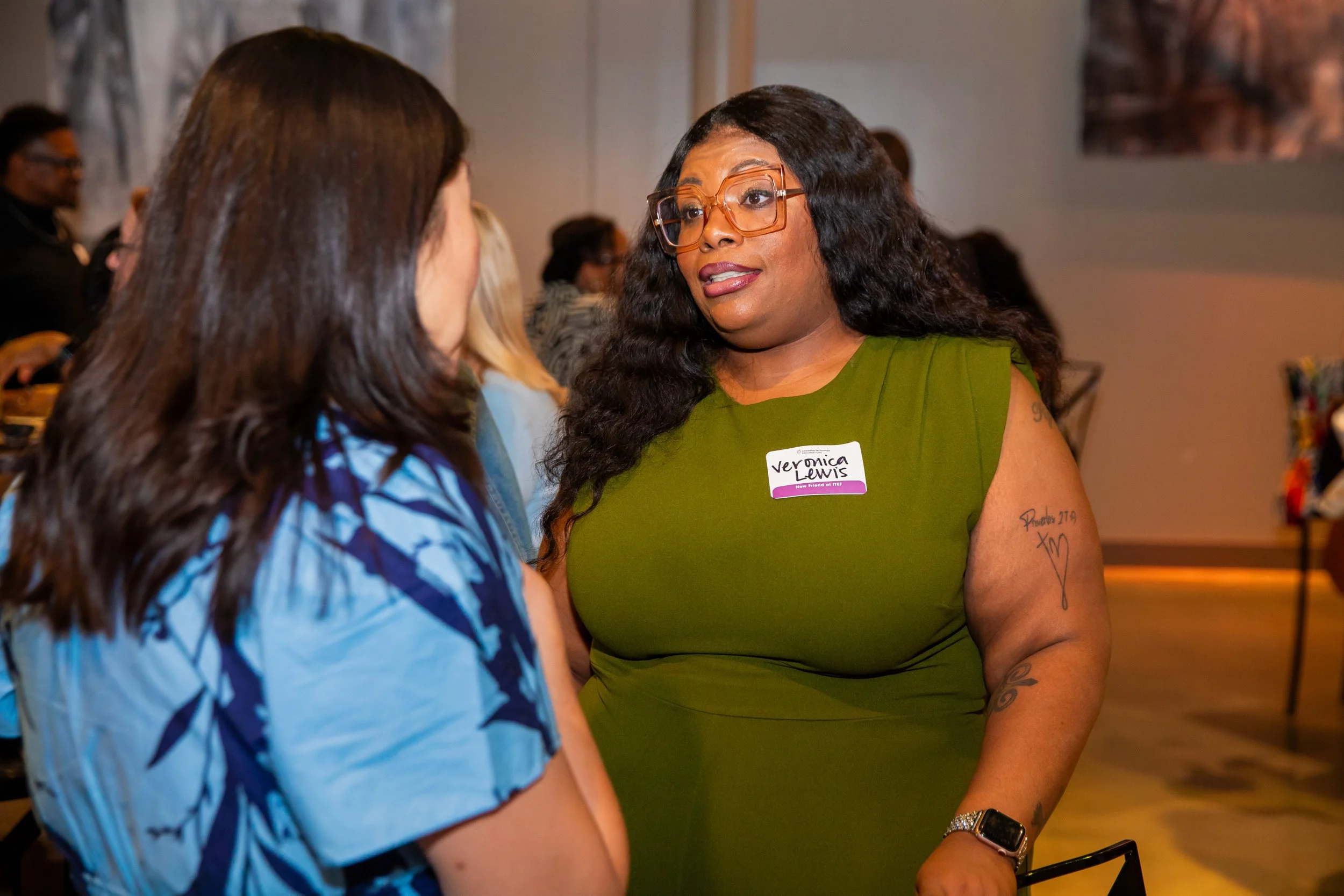 A woman with long curly hair wearing glasses and a green dress is talking to another woman with long brown hair wearing a blue patterned top at an indoor event.