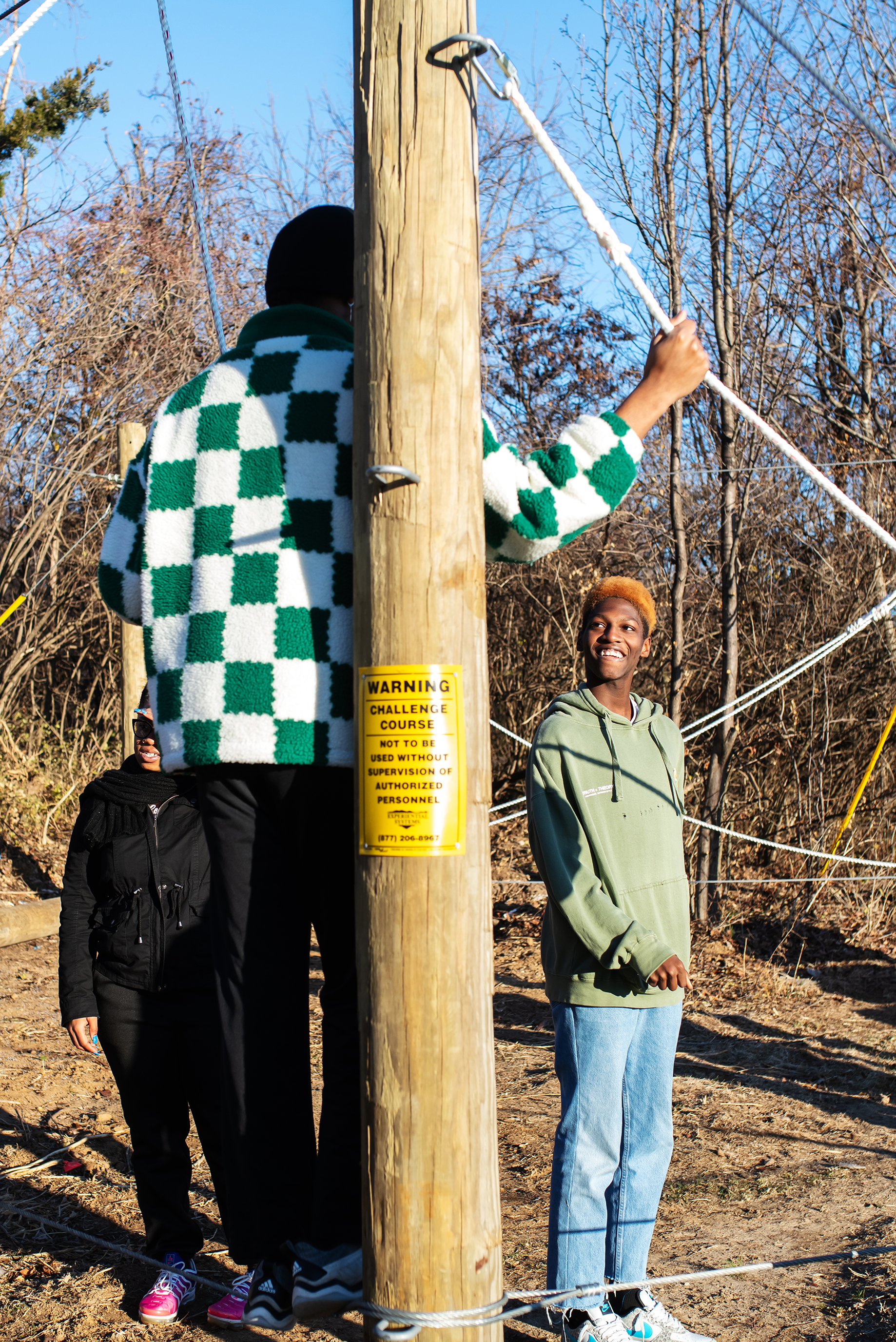 People standing outdoors near a wooden pole with a yellow warning sign, among leafless trees under a clear blue sky.