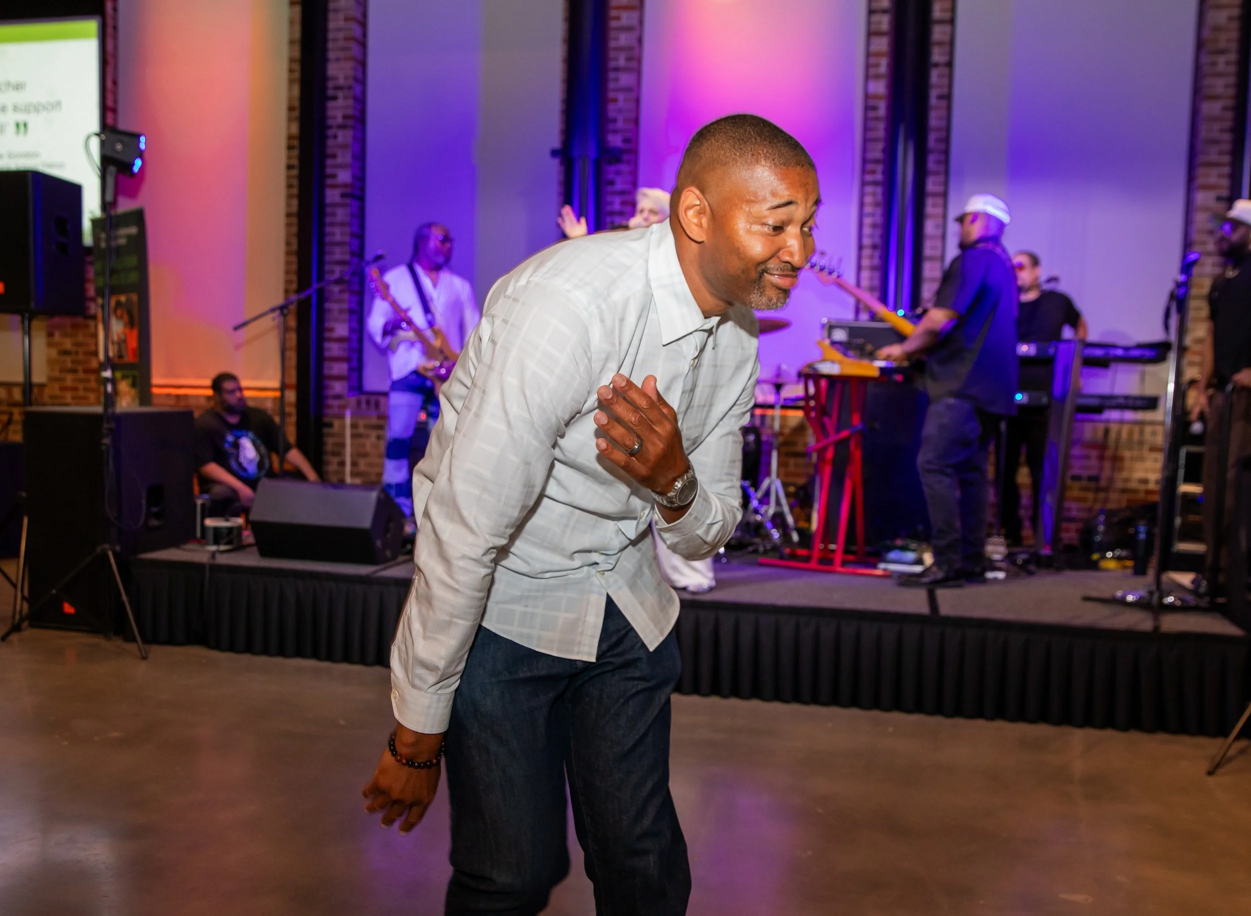 A man dancing with a smile, wearing a white checked shirt and dark jeans, in a lively indoor event with a band performing in the background, purple and orange stage lighting.