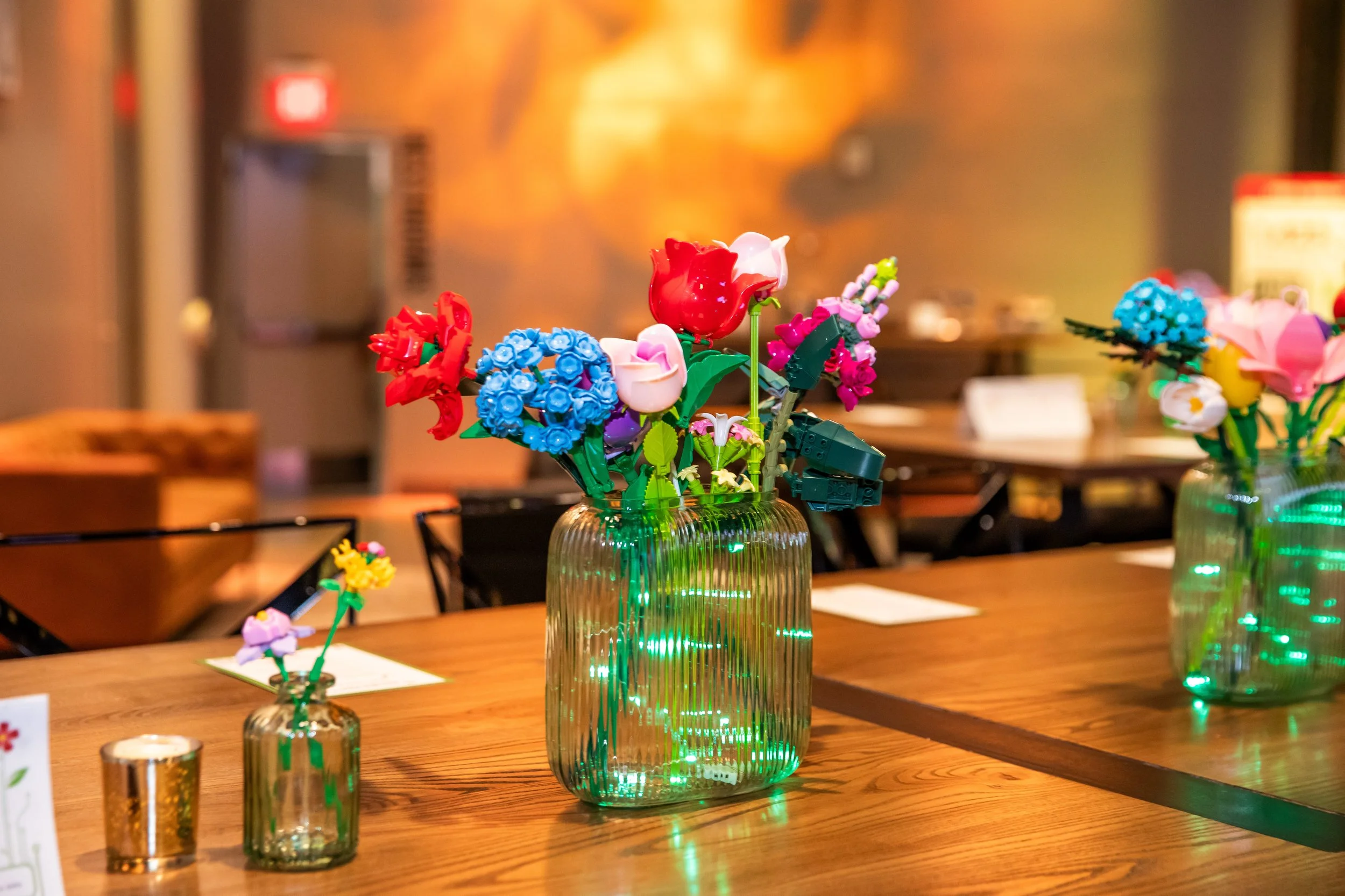 A clear glass vase with vertical ridges filled with artificial colorful flowers, including red, white, pink, blue, purple, and yellow, placed on a wooden table.