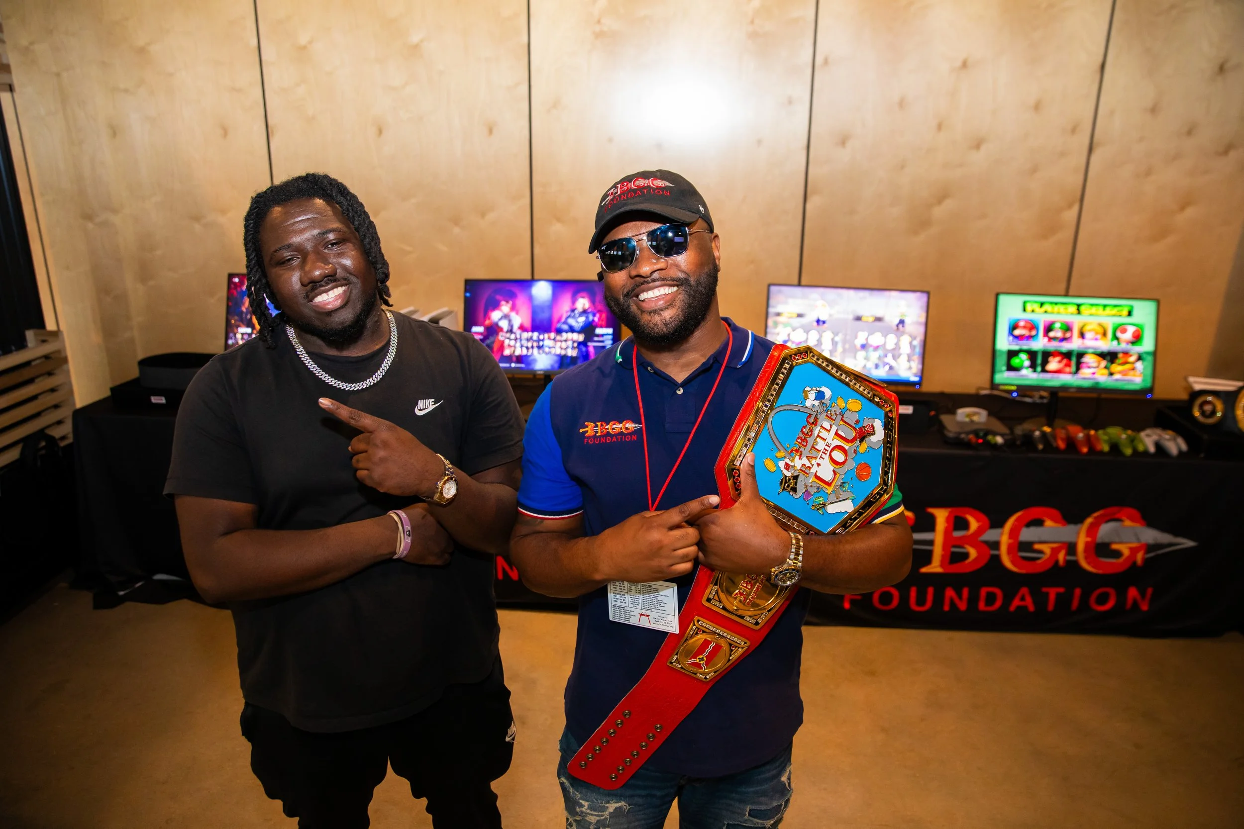 Two men smiling, one pointing at the other, in front of a display with multiple monitors, at an event for the Bob G Foundation, with a man holding a championship belt.