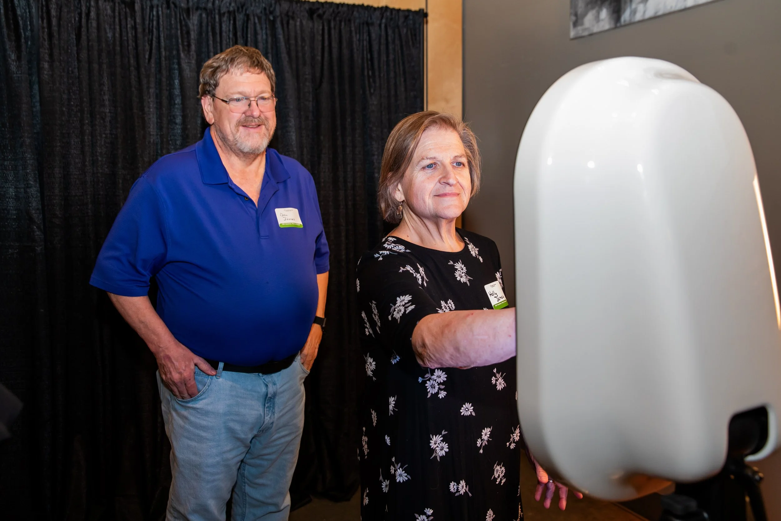 A man with glasses and a beard wearing a blue polo shirt and jeans stands next to a woman with short hair wearing a black dress with white floral patterns. She is operating a large, white, oval-shaped machine, possibly a 3D scanner or a medical imagi