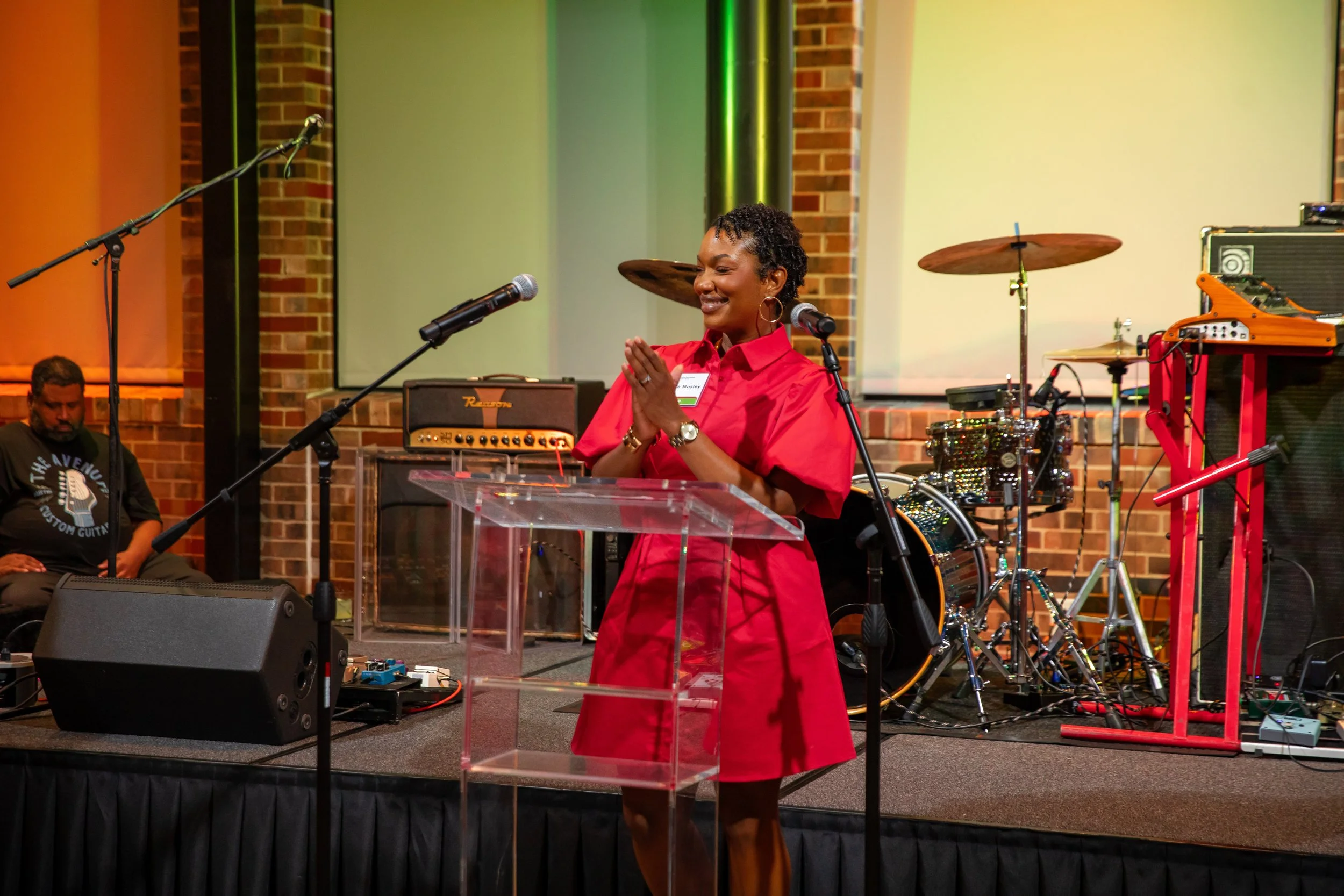 A woman in a red dress standing at a clear podium with her hands clasped, smiling with eyes closed. She is in front of a microphone, with musical instruments and sound equipment behind her on a stage with brick walls and a green screen.