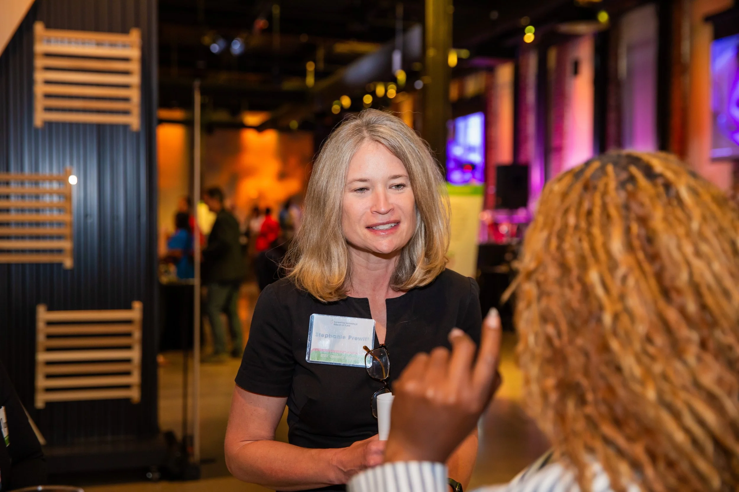 A woman with blonde hair wearing a black top and glasses hanging from her neckline at a social event, engaged in conversation with another person with curly hair. The woman has a name tag that reads 'Stephanie Prewitt'.