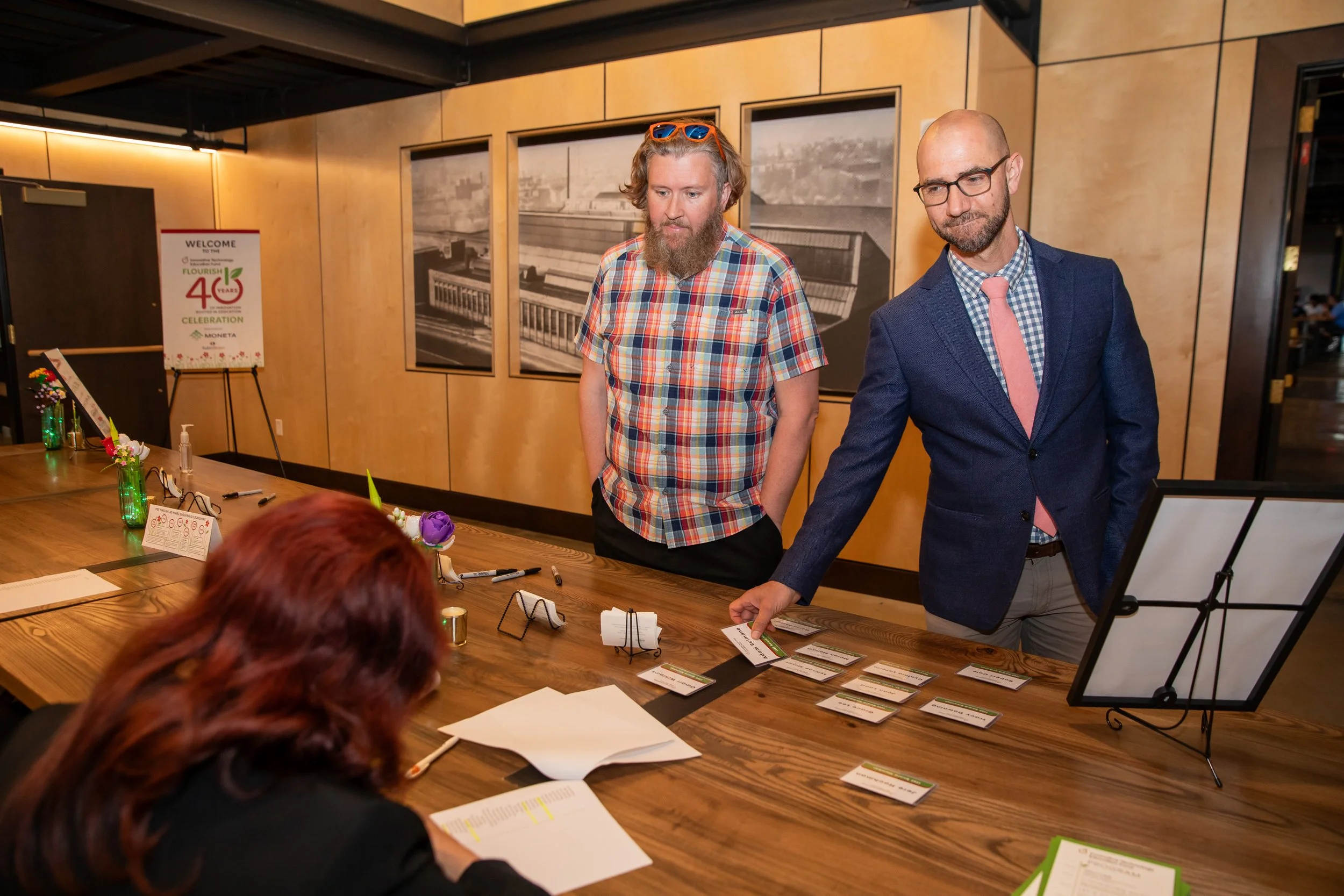 Two men are standing at a conference table in a modern office setting. One man is wearing a suit with a pink tie and glasses, while the other is dressed casually in a colorful plaid shirt with sunglasses on his head. There are papers, name tags, and 
