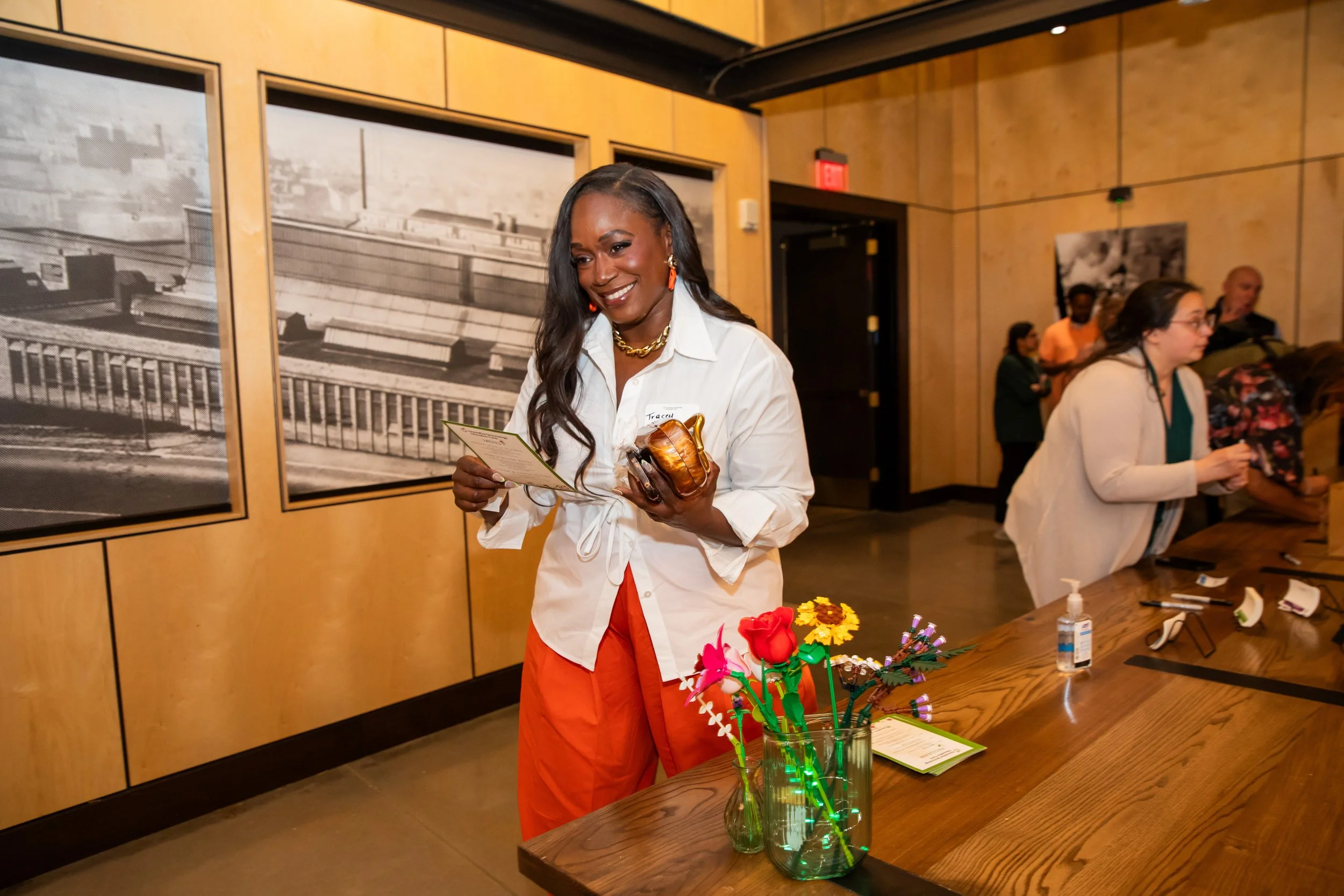 A woman with dark hair wearing a white shirt and orange pants is smiling while holding a certificate and a small pumpkin-shaped object. Other people are in the background at an indoor event with wooden walls and black-and-white photographs.