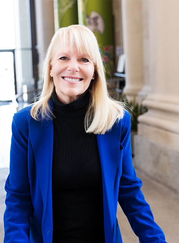 A smiling blonde woman wearing a black turtleneck and a blue blazer, standing indoors near a column with a green wall and windows behind her.