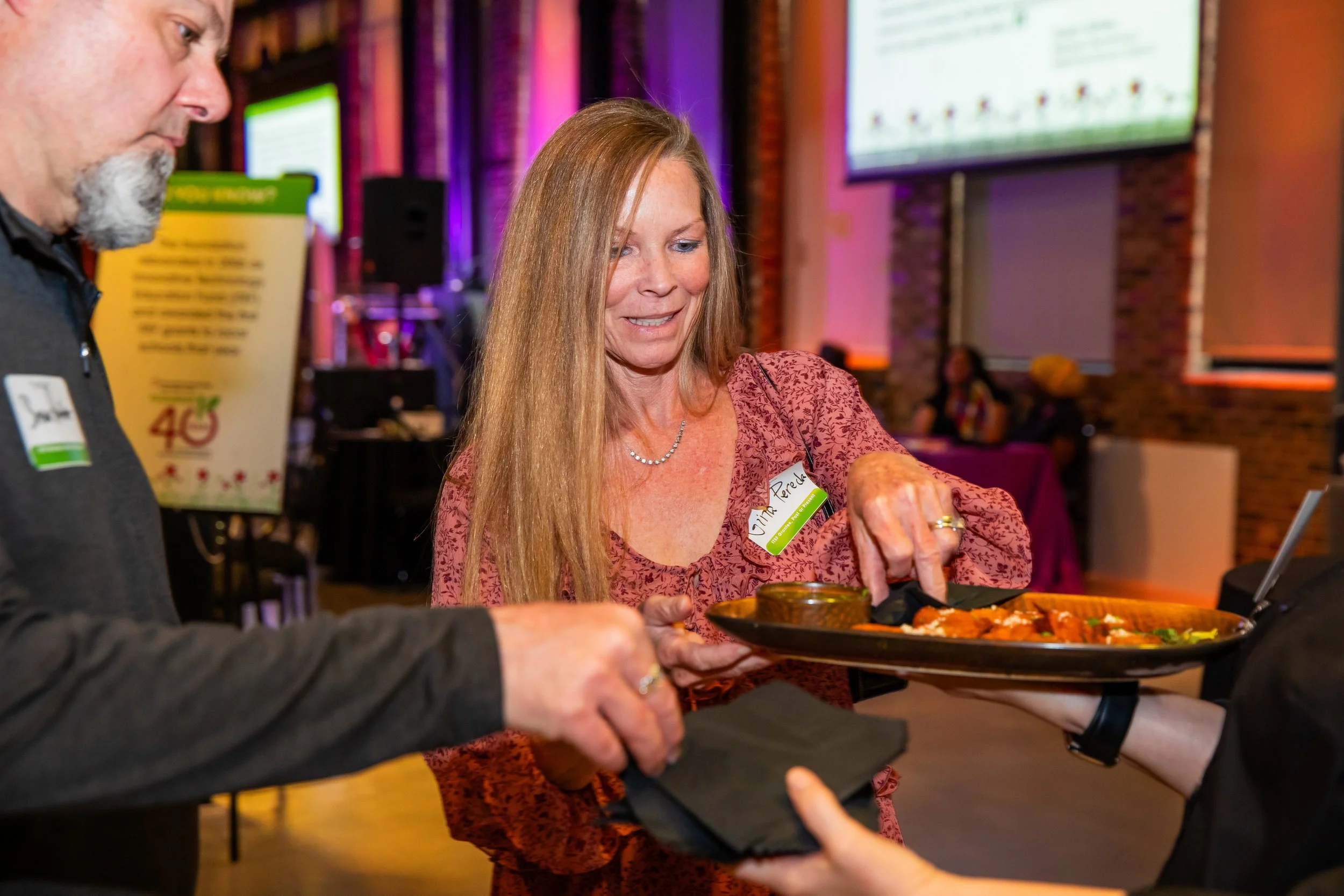 A woman with long red hair, wearing a pink dress and a pearl necklace, is serving food onto a plate at an indoor event. She is smiling and interacting with others who are also serving or receiving food. The background shows a brightly lit room with l