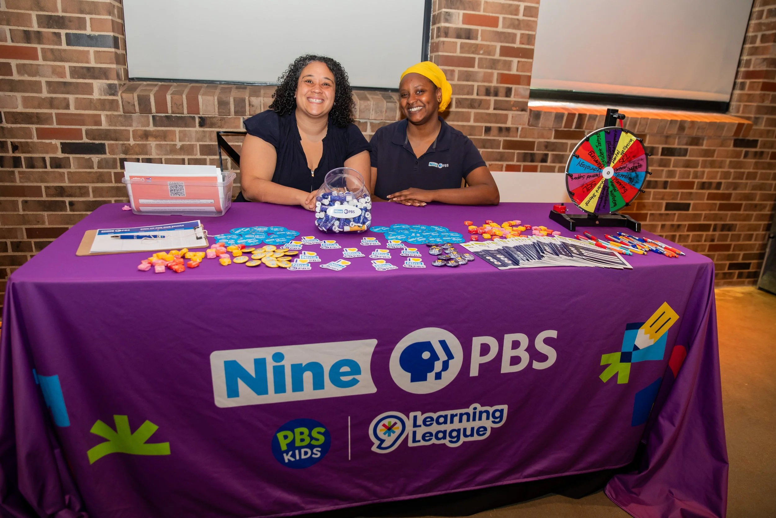 Two women sitting behind a table with promotional materials, a prize wheel, and a clear jar filled with blue and white items. The table is covered with a purple cloth displaying logos for Nine PBS, PBS Kids, and Learning League. They are smiling, wit