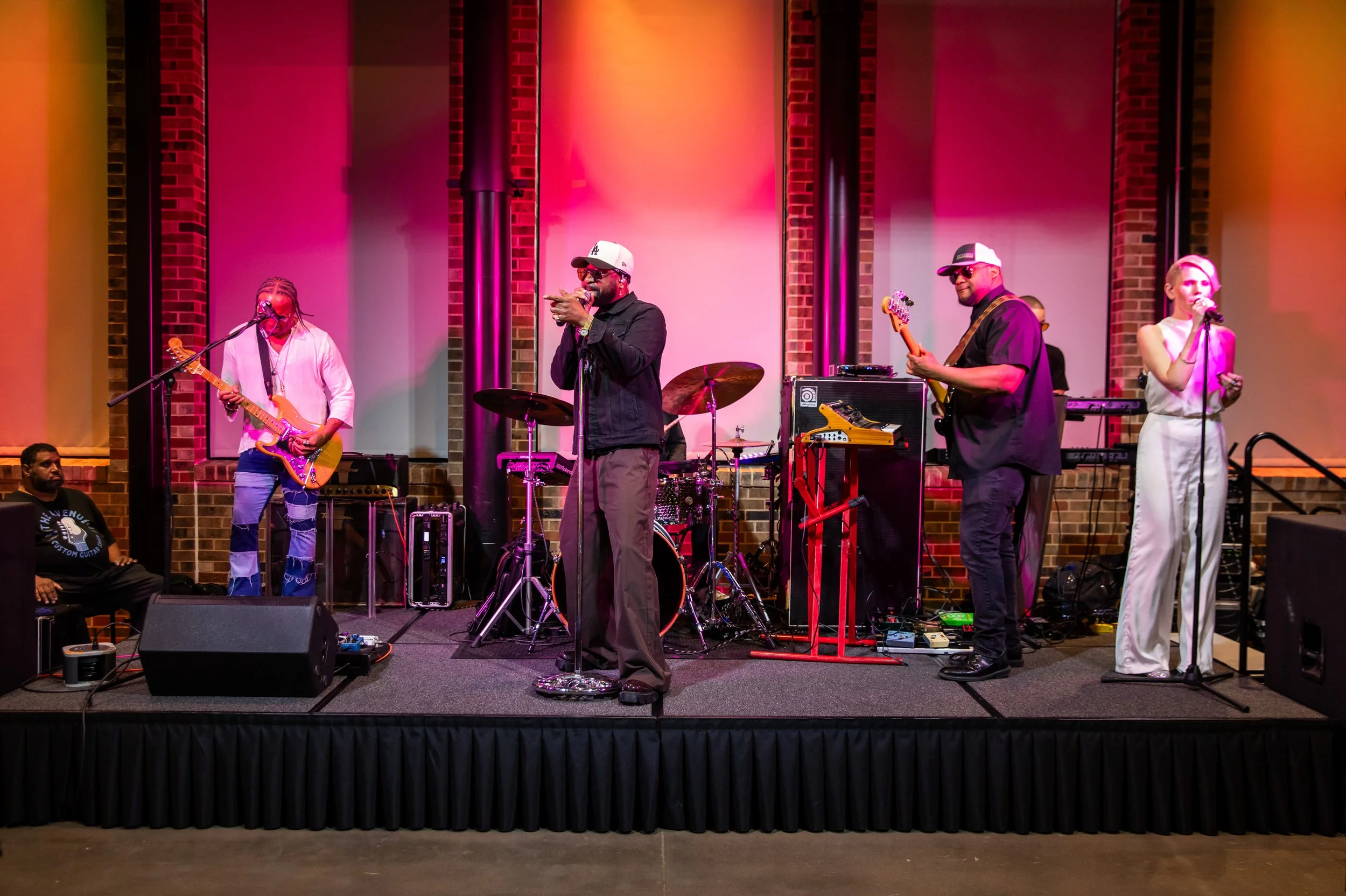 A band performing on stage with multiple musicians including a guitarist, a singer, and a bassist, inside a venue with brick walls and pink lighting.