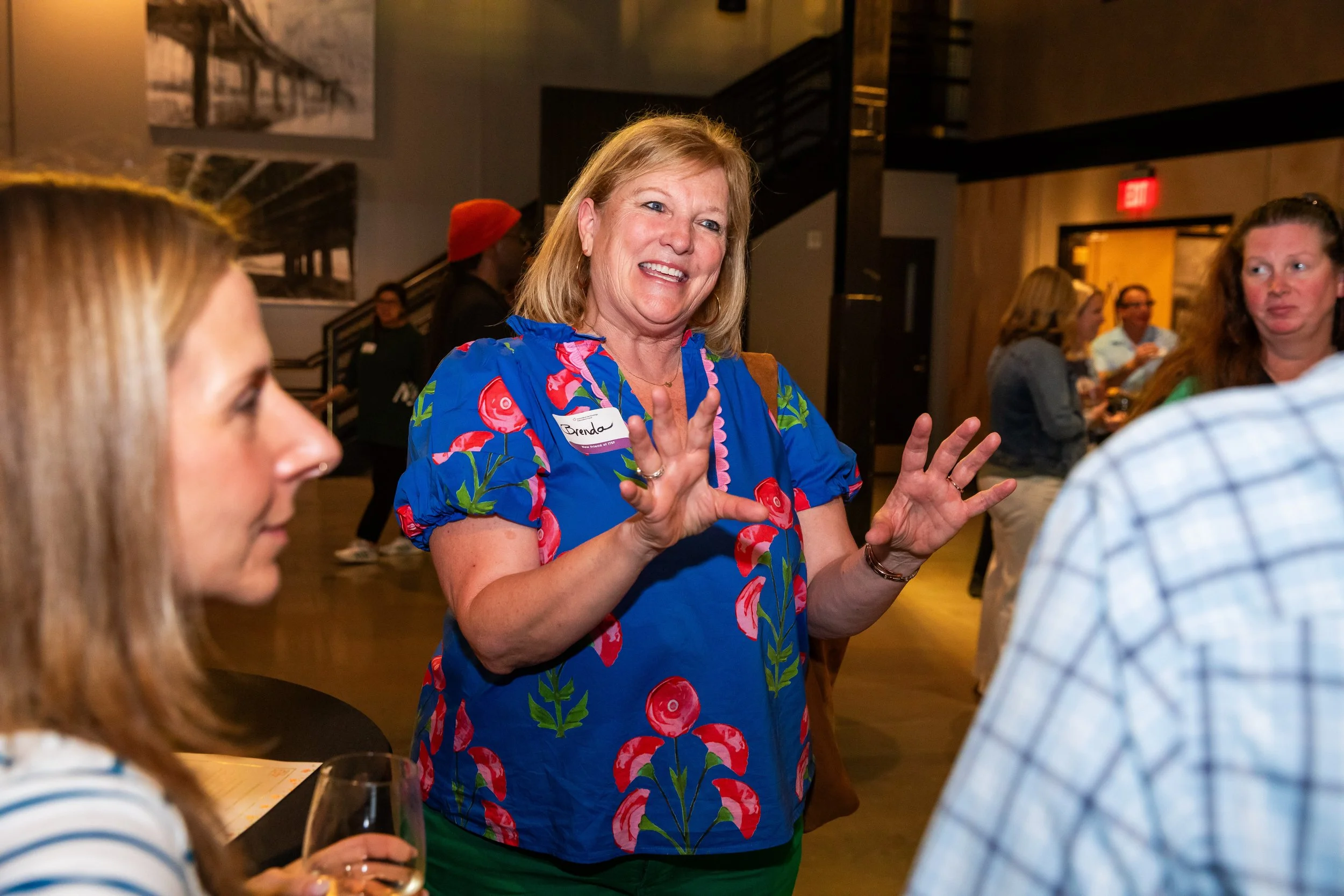 A woman with a name tag that reads 'Brenda' is smiling and gesturing with her hands while talking to others at a social gathering. She has blonde hair and is wearing a colorful blue dress with a cherry pattern. Several other people are visible in the
