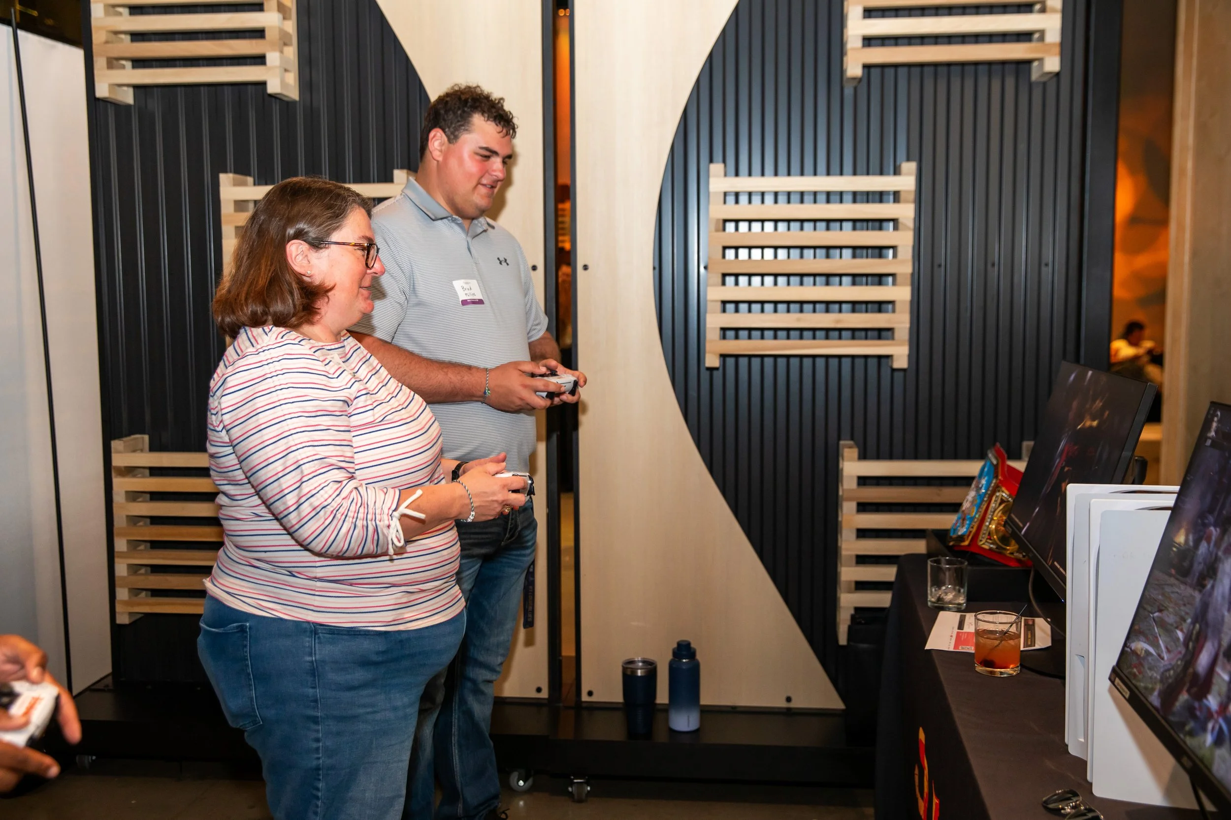 Two people, a woman and a man, are standing in front of a computer monitor at an indoor event. The woman is wearing a striped shirt and glasses, and the man is wearing a gray polo shirt and holding a gaming controller.